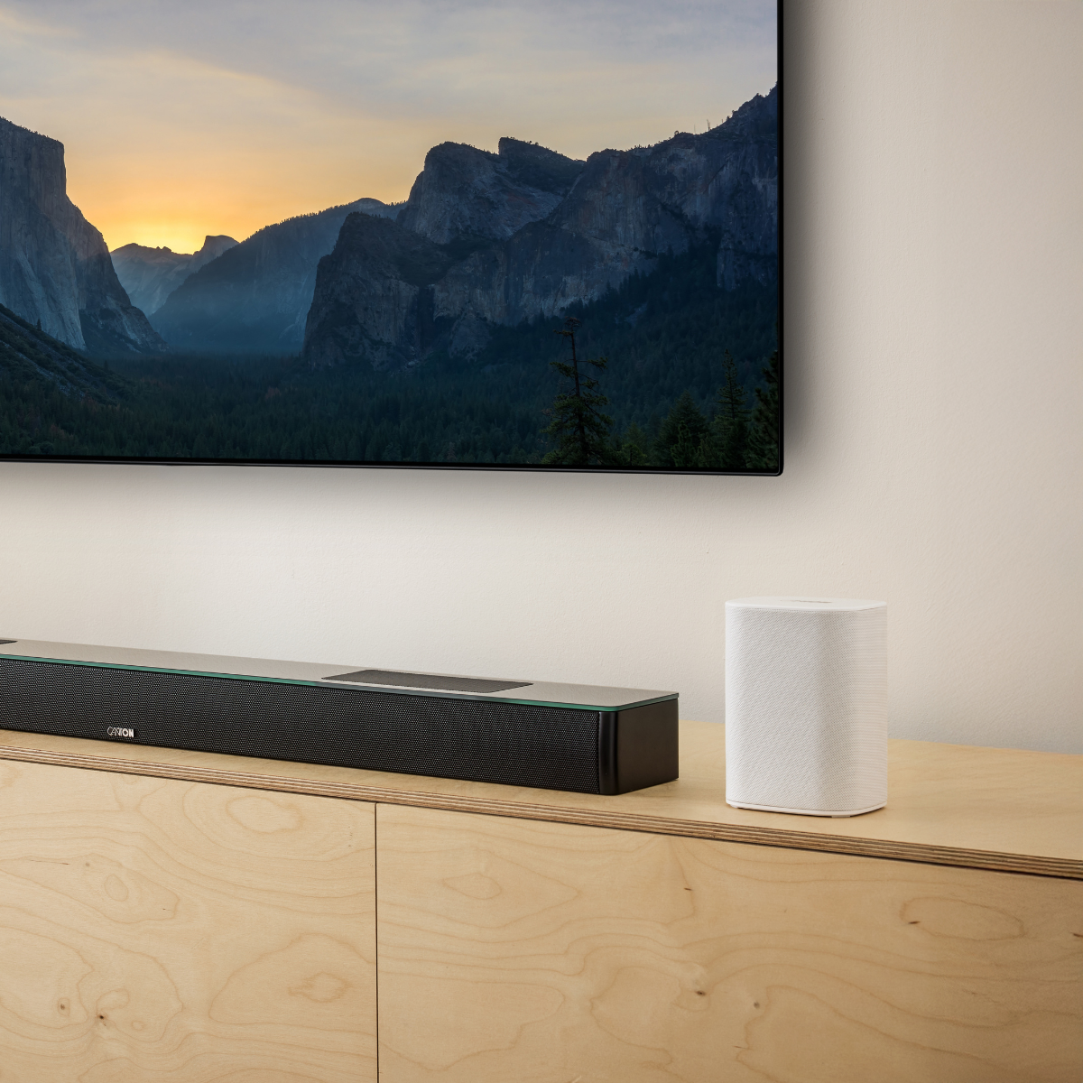 Ein schwarzer Soundbar und ein weißer Lautsprecher stehen auf einem Holzschrank vor einem Bergpanorama.