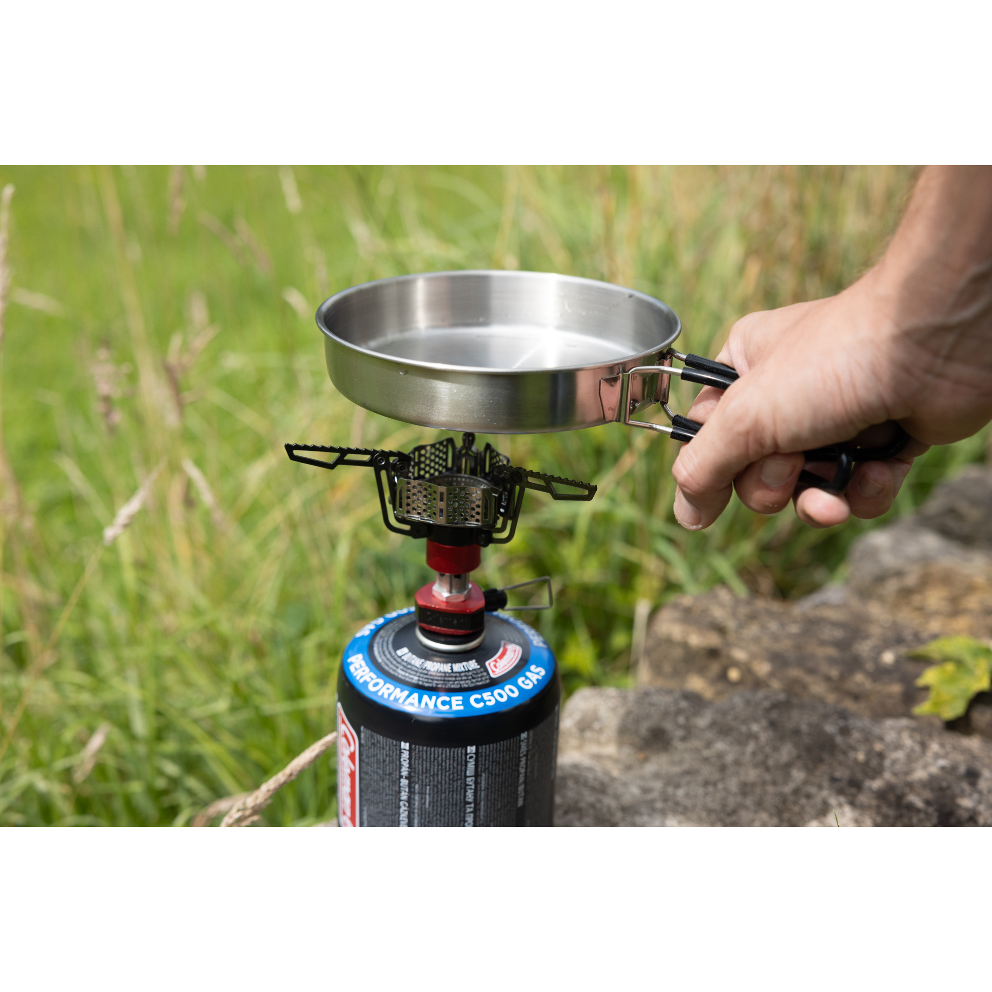 Eine Hand hält eine Pfanne über einem Campingkocher mit Propangasflasche.