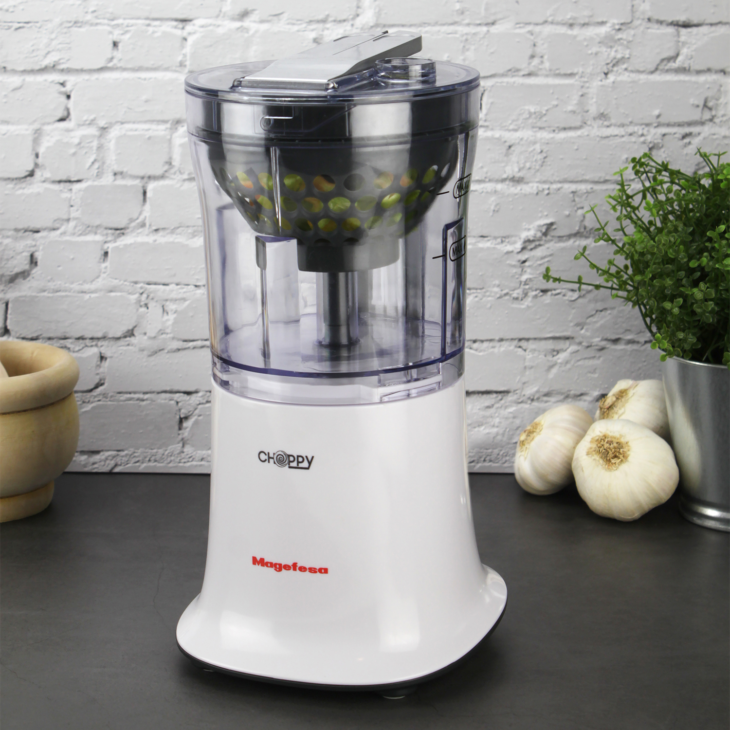 Un picador de alimentos Magefesa Choppy blanco con recipiente transparente en encimera oscura.