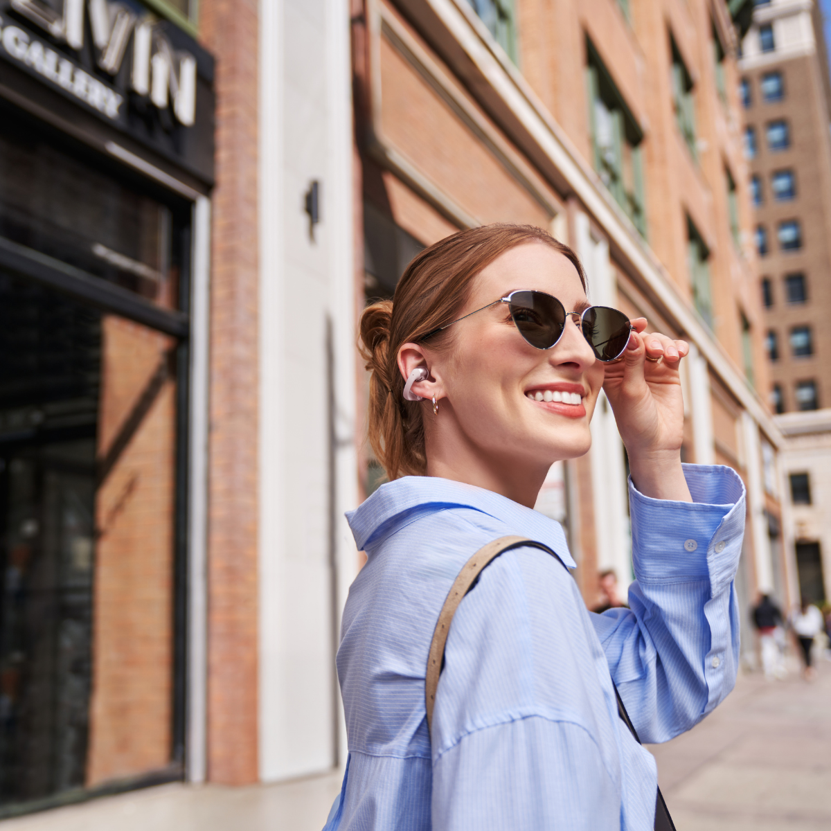 A woman with earbuds and sunglasses smiles, looking aside, in front of a brick building.
