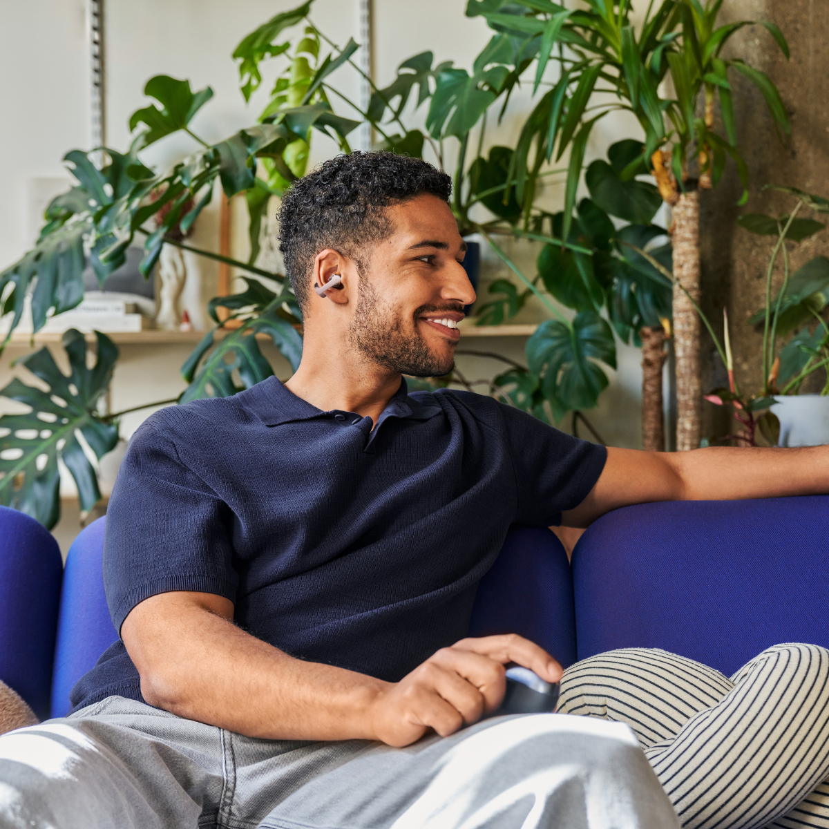Man with earbuds on a blue couch, surrounded by plants.