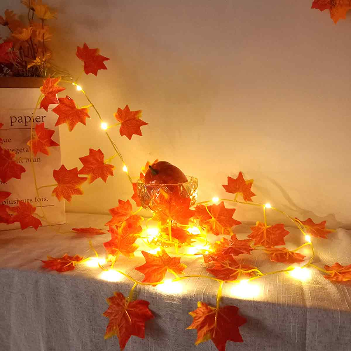 Herbstlaub-Lichterkette mit roten Ahornblättern und einer kleinen Schale mit Apfel.