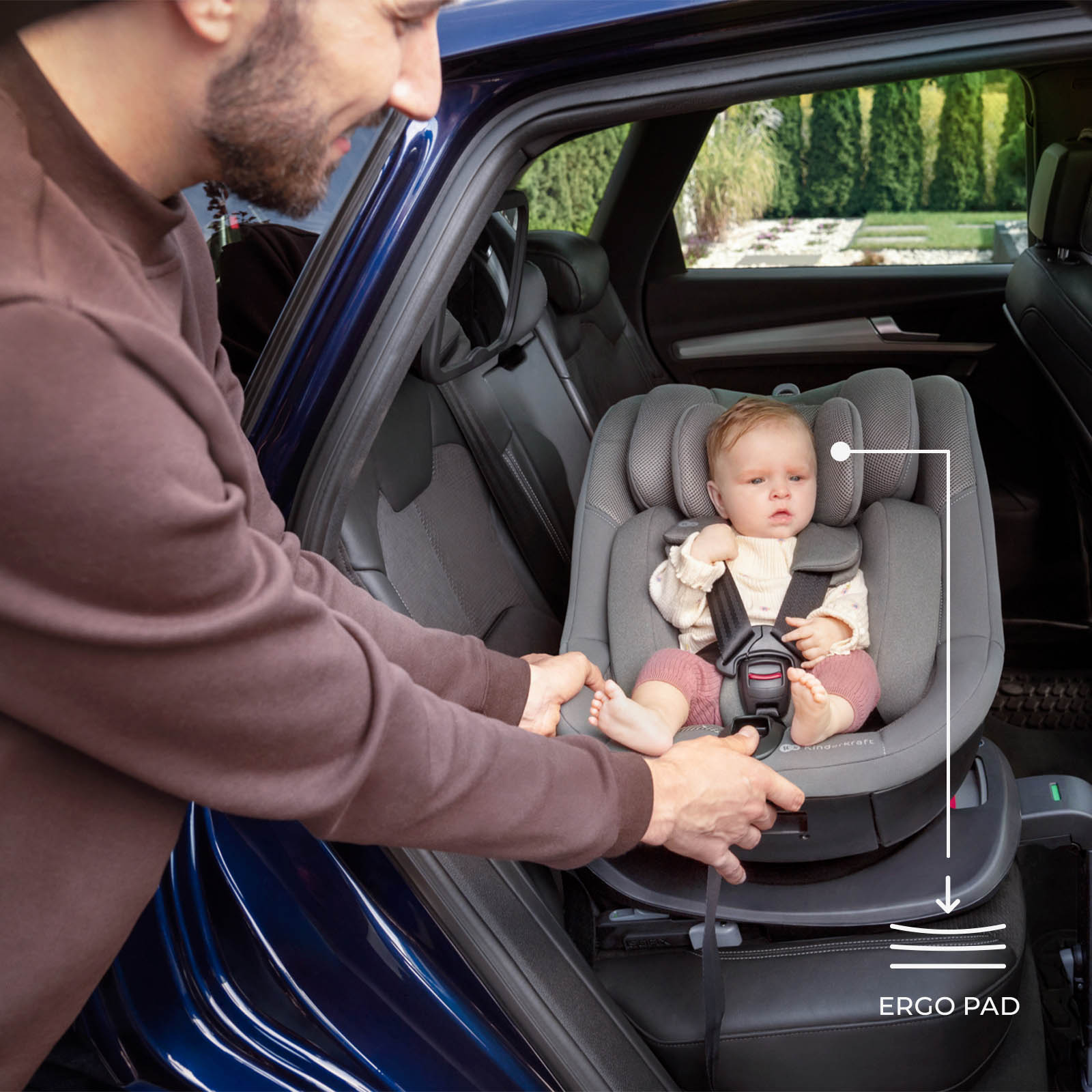 Ein Mann befestigt einen Kindersitz mit Baby im Auto. Draußen ist eine grüne Landschaft.