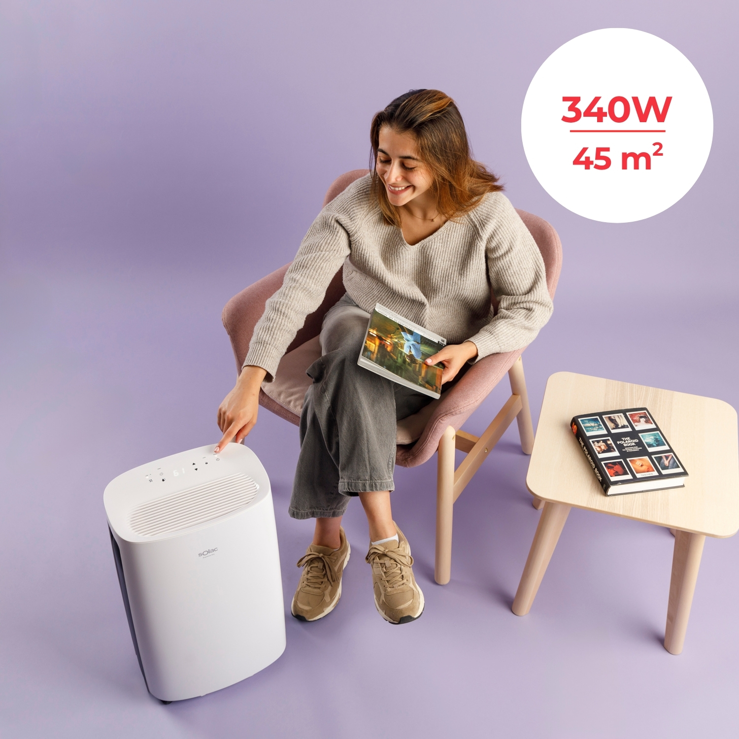 Mujer en silla toca purificador de aire. Libro en mesa. Info 340W 45m².