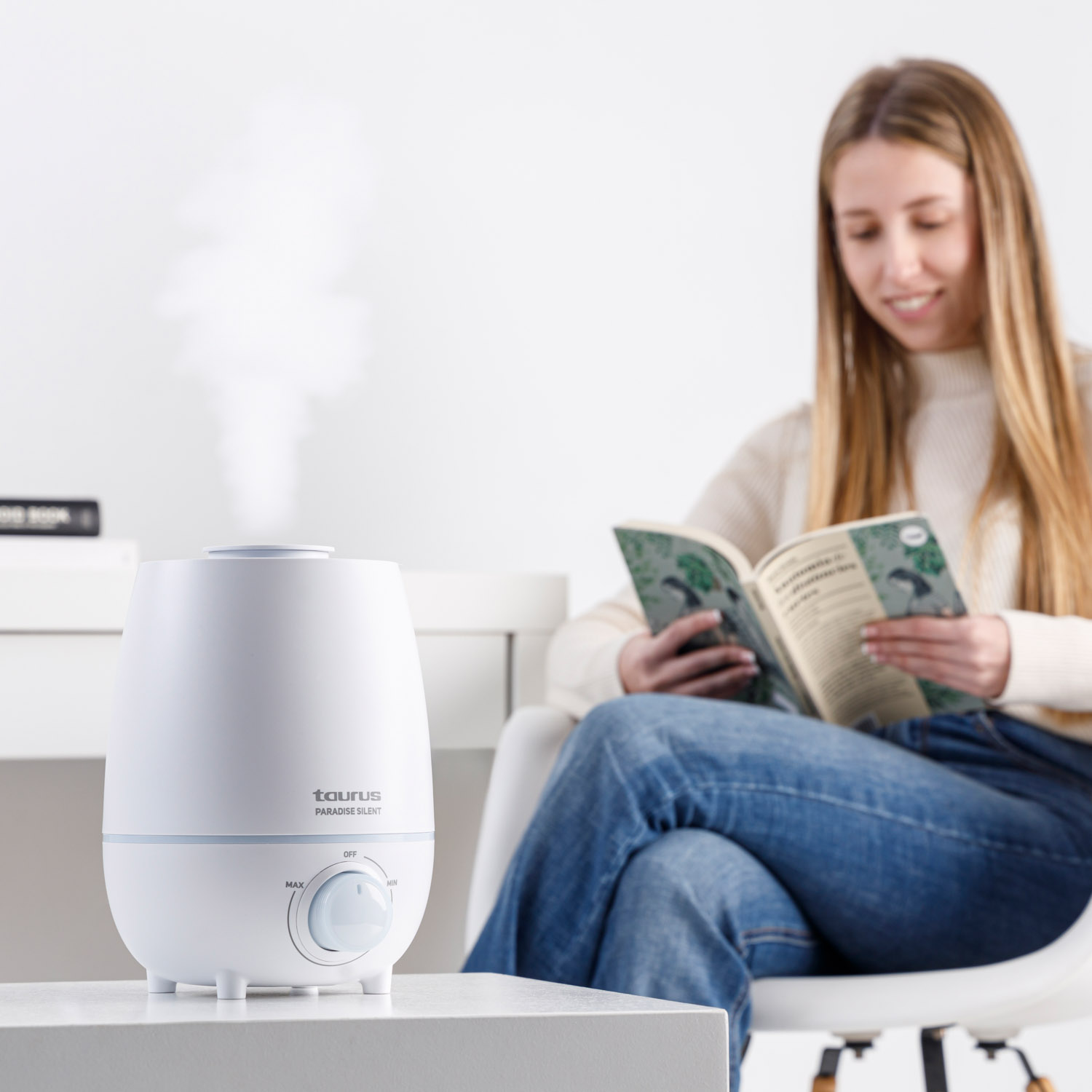 Un humidificador blanco emite vapor en una mesa, con una mujer leyendo al fondo.