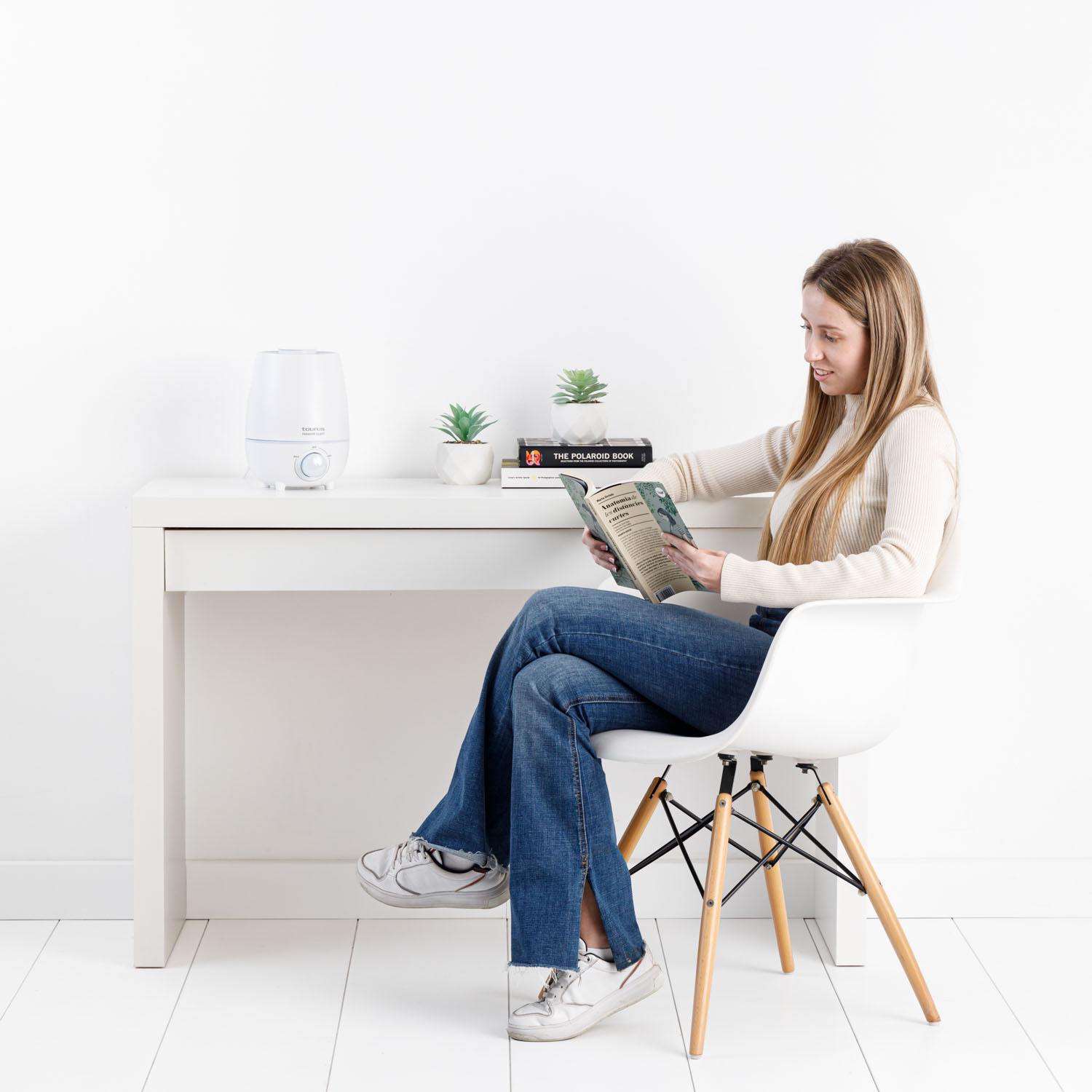 Mujer leyendo libro en escritorio con humidificador y plantas.