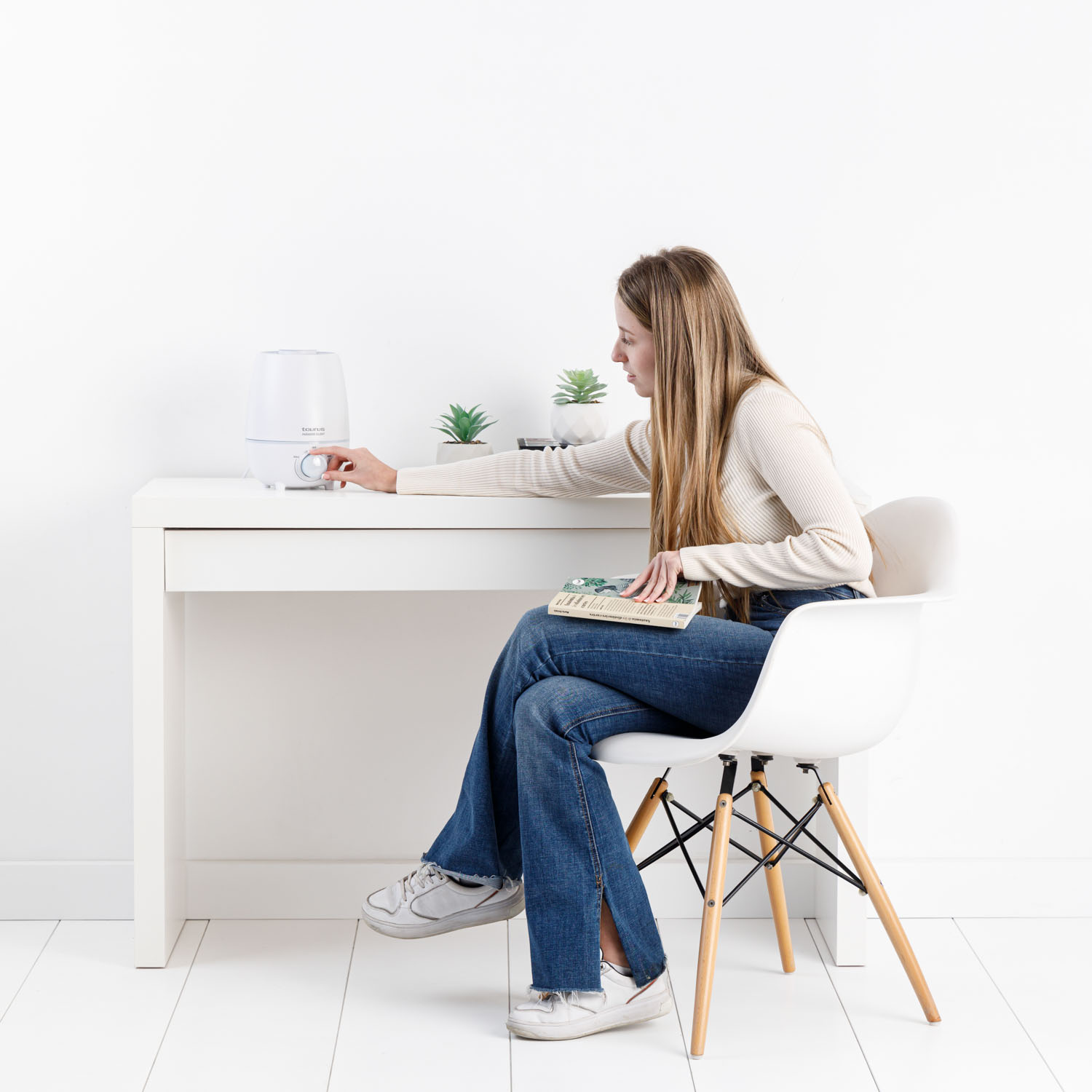 Una mujer se sienta en un escritorio, ajustando un humidificador blanco. Dos plantas en maceta están sobre el escritorio.