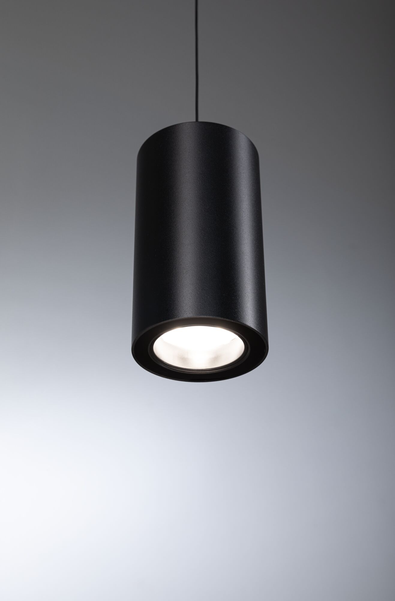 Eine schwarze, zylindrische Pendelleuchte leuchtet hell vor einem grauen Hintergrund.
