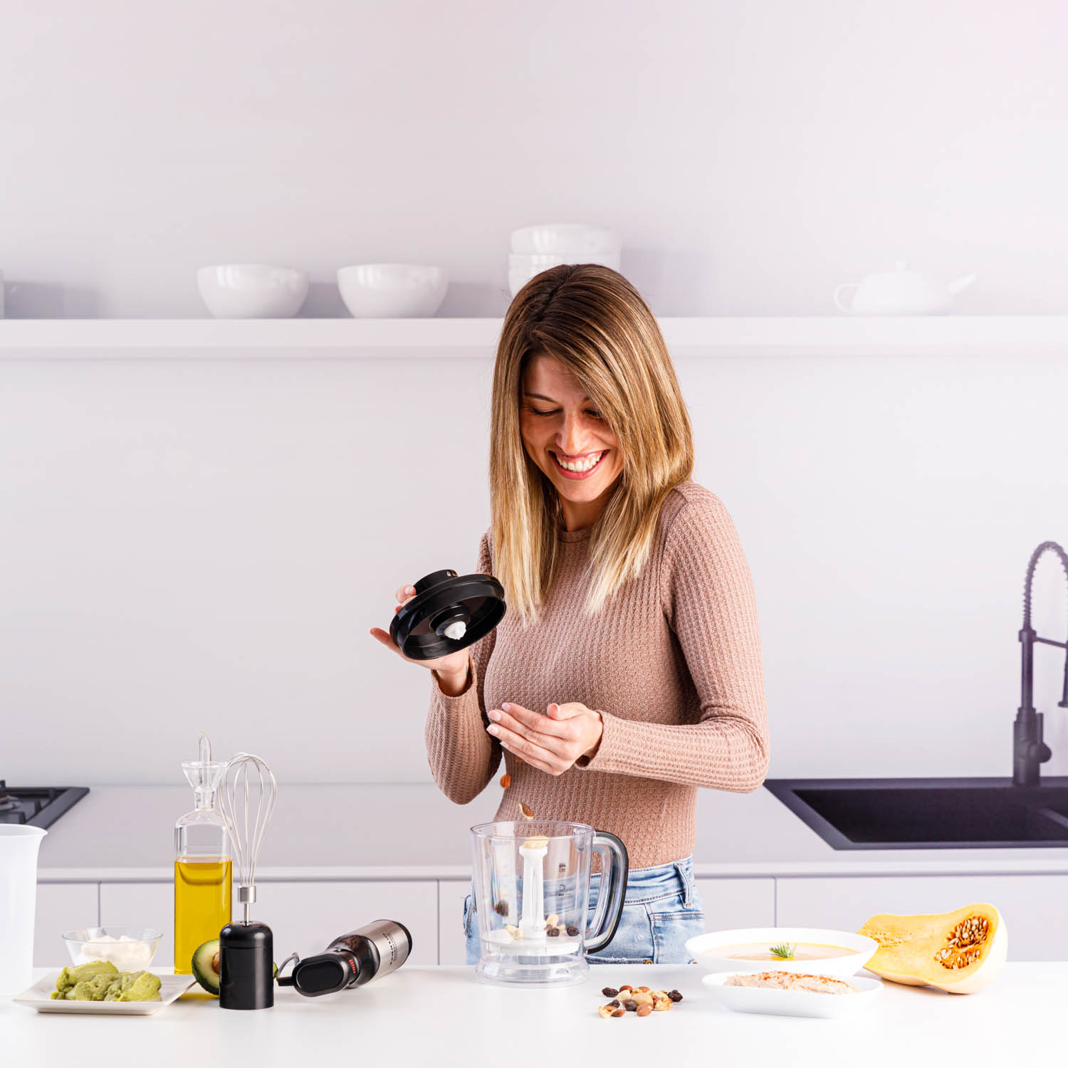 Una mujer en la cocina sostiene la tapa de una licuadora. Hay ingredientes en la encimera.