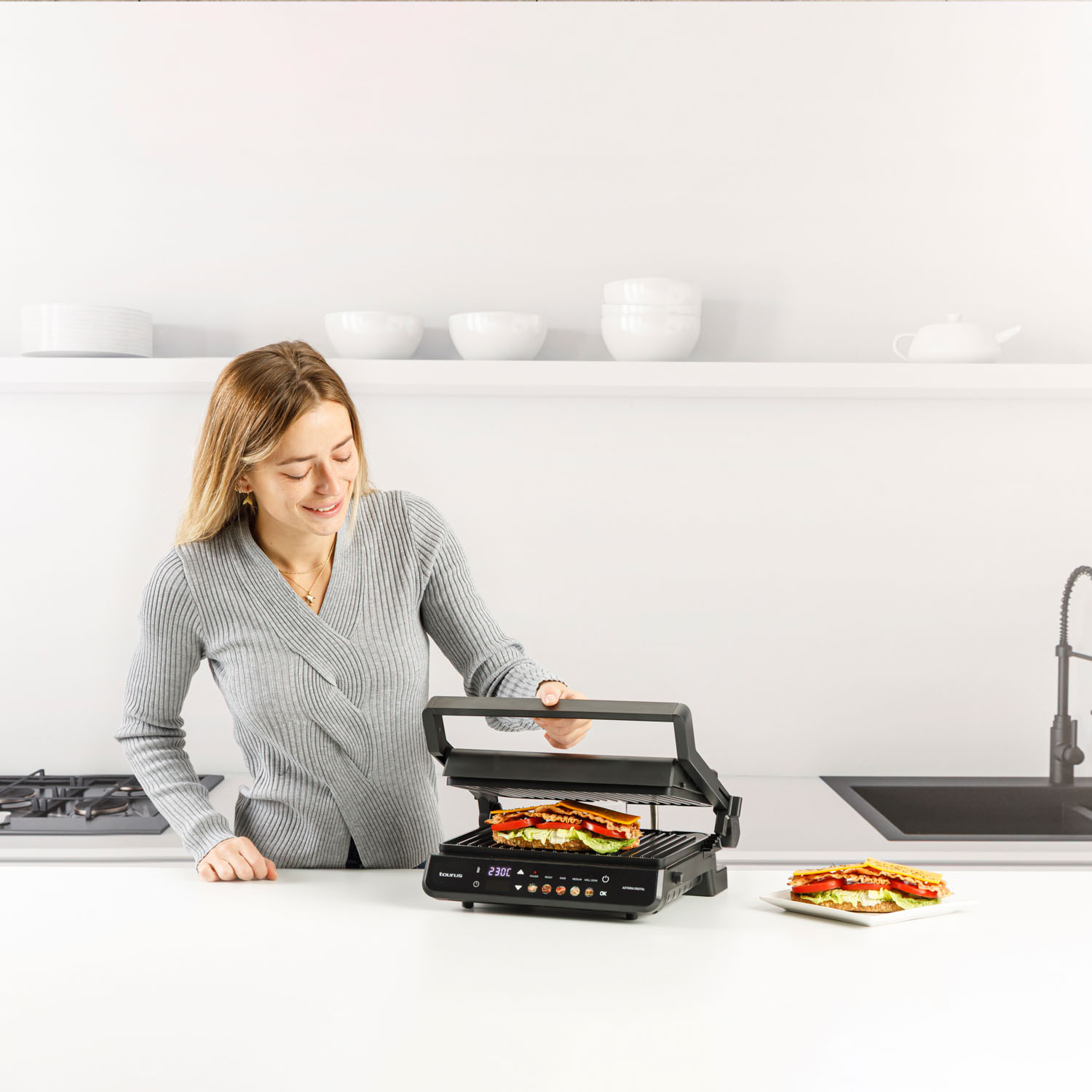 Una mujer con un suéter gris está en una cocina, mirando una sandwichera.