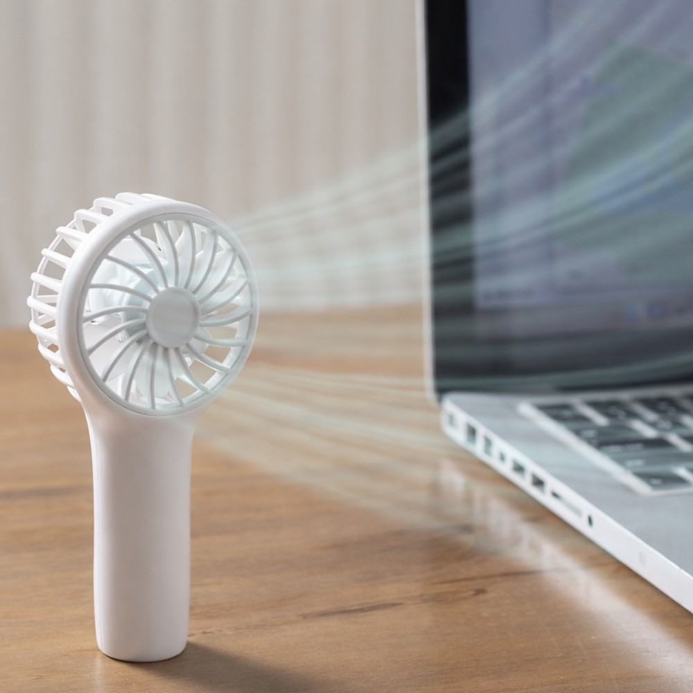 Un ventilador blanco de mano sopla aire hacia una laptop abierta en un escritorio de madera.
