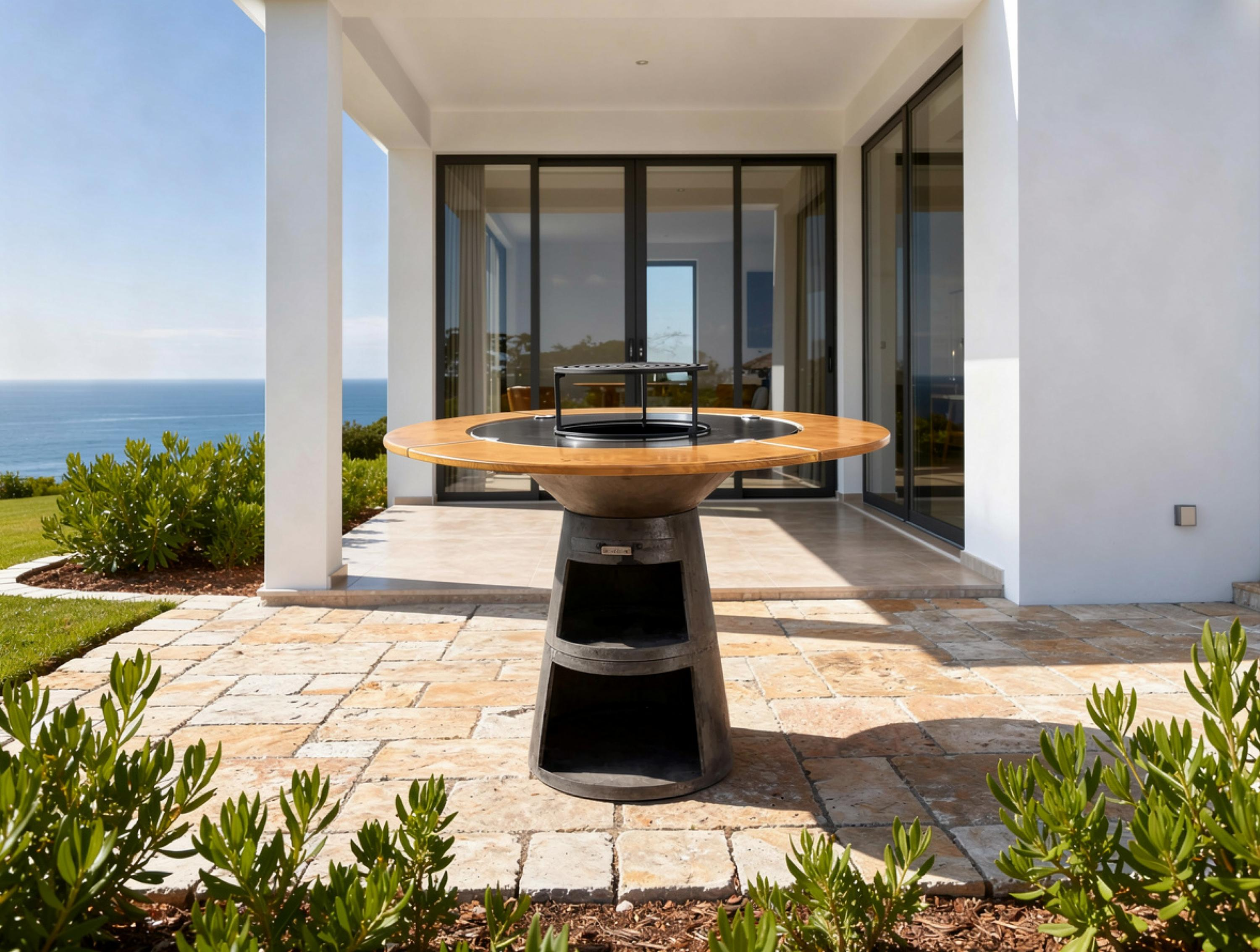 Moderner Grilltisch aus Metall mit Holzplatte vor einem Haus am Meer.