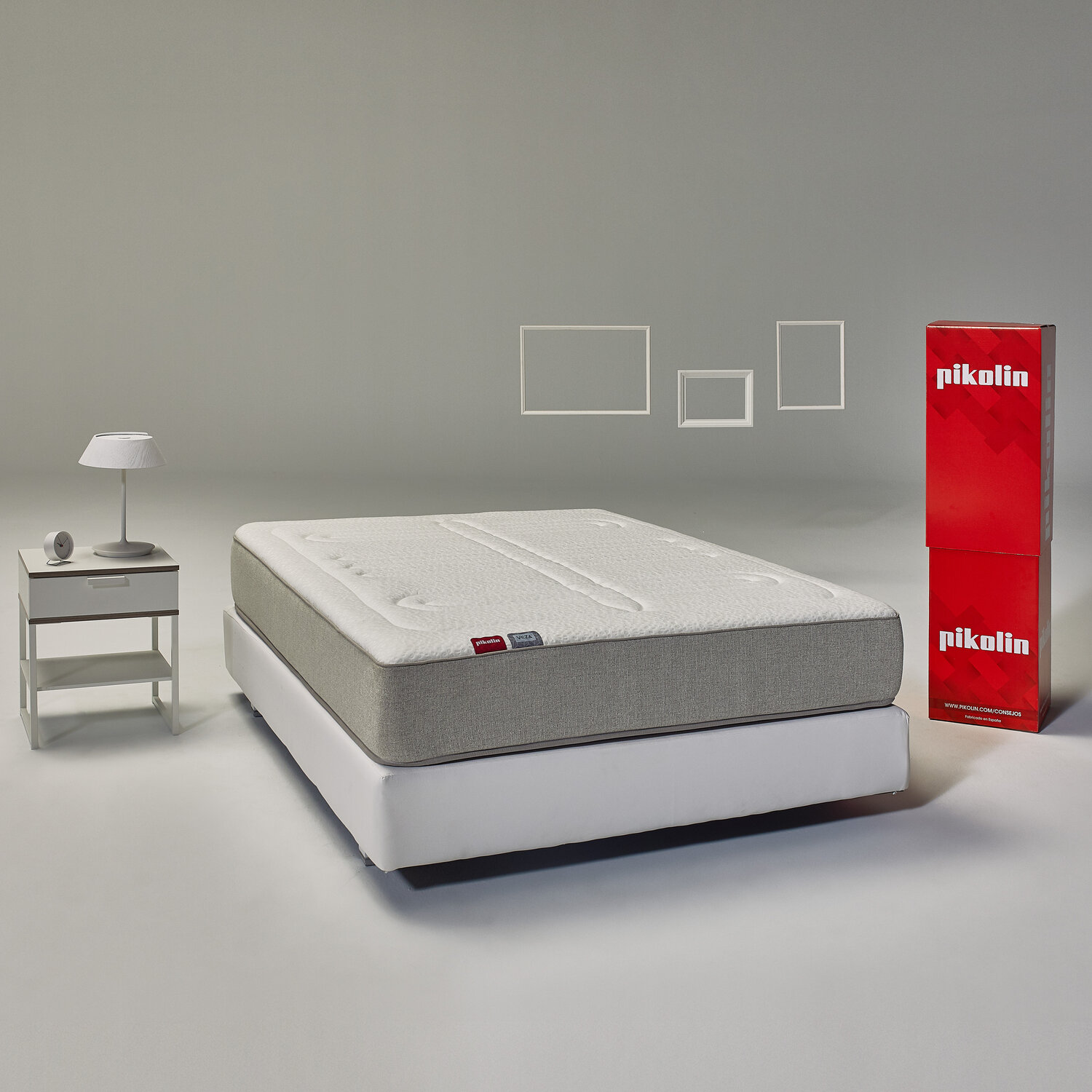 Cama con base blanca, lateral gris y tapa blanca. Mesita de noche, lámpara, reloj y cajas rojas.