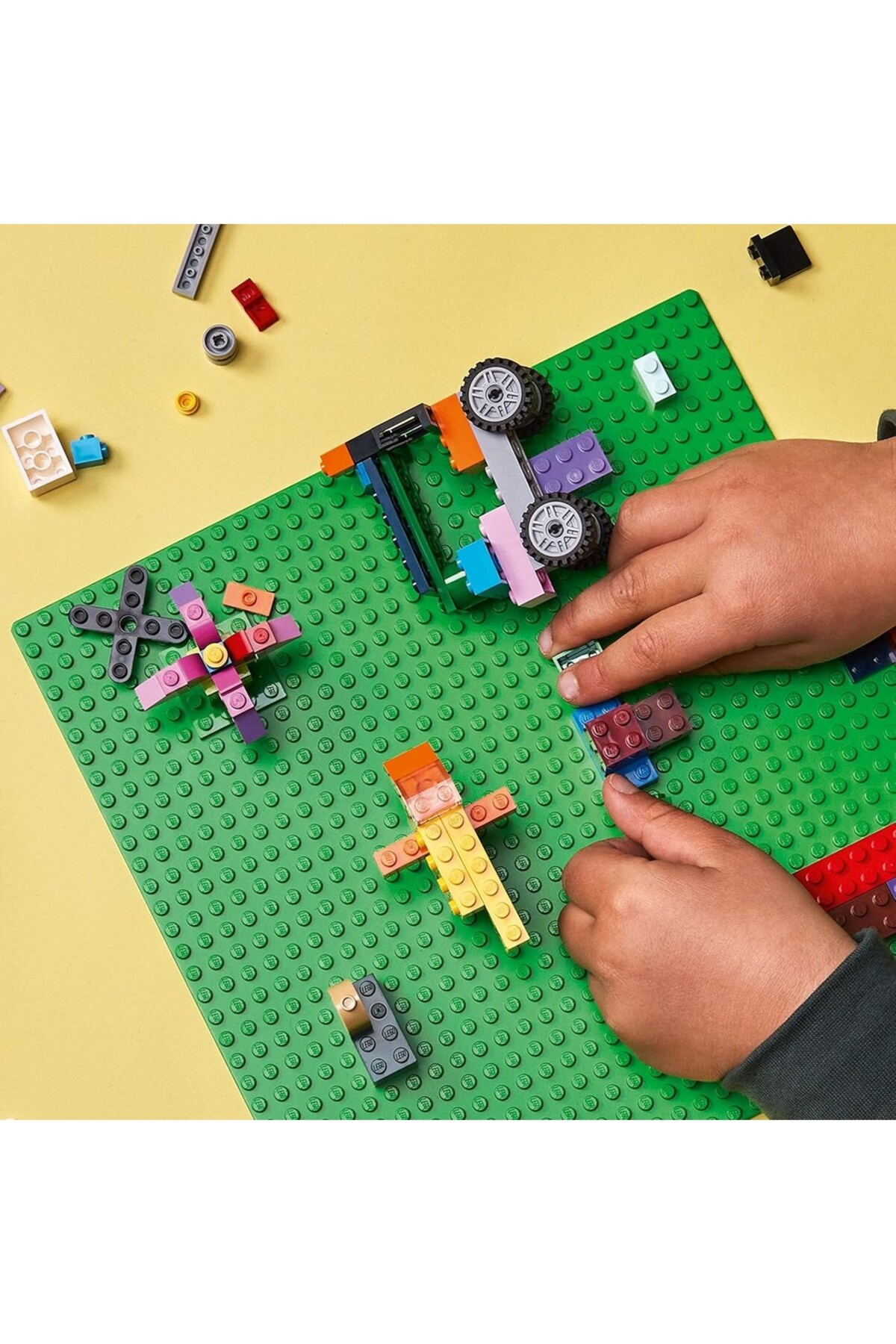 Çocuklar yeşil bir zeminde renkli Lego tuğlalarıyla inşa ediyor. Araba ve figürler yapılıyor.