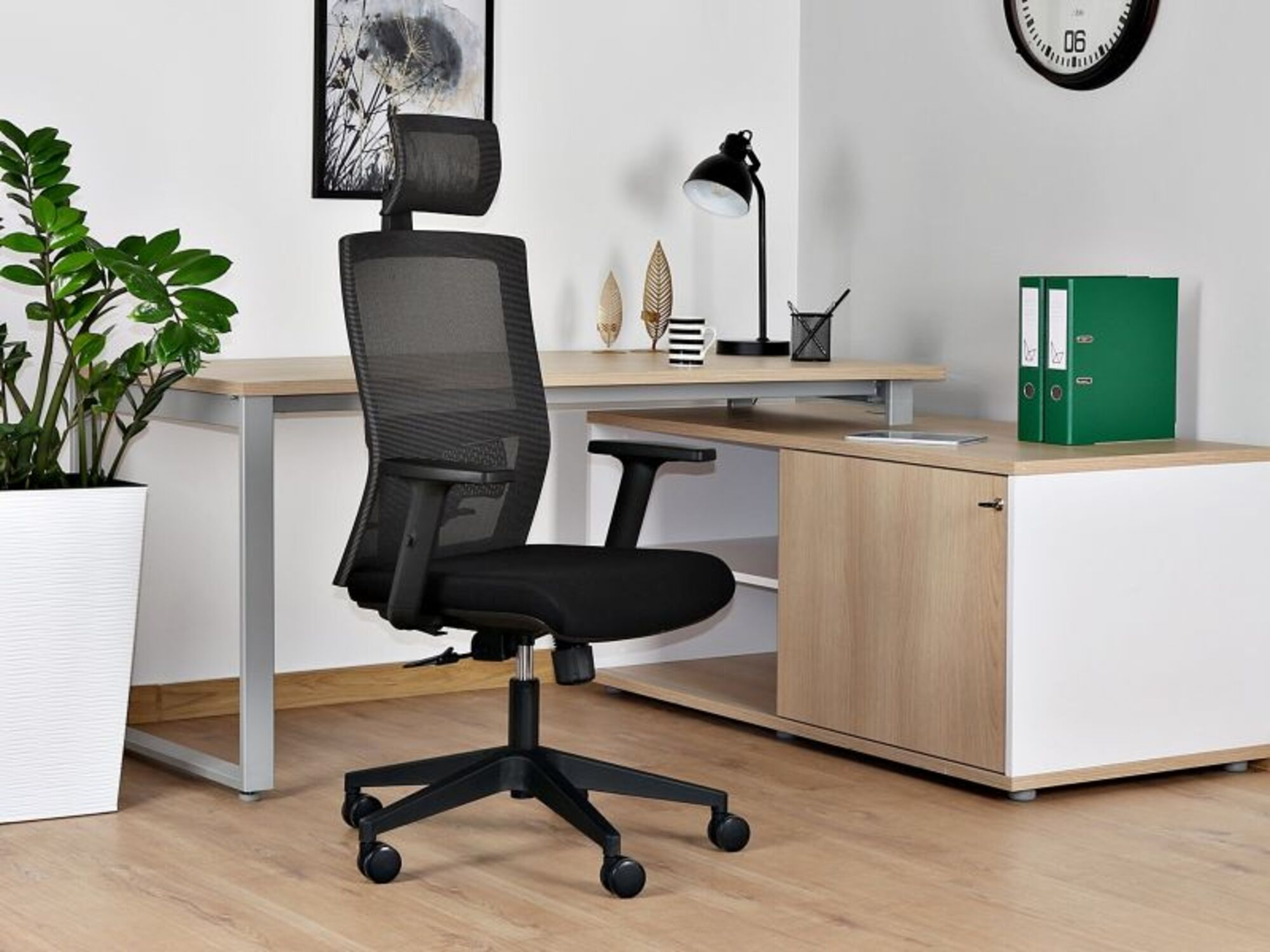 Czarny fotel biurowy przed biurkiem z roślinami, lampą i segregatorami.