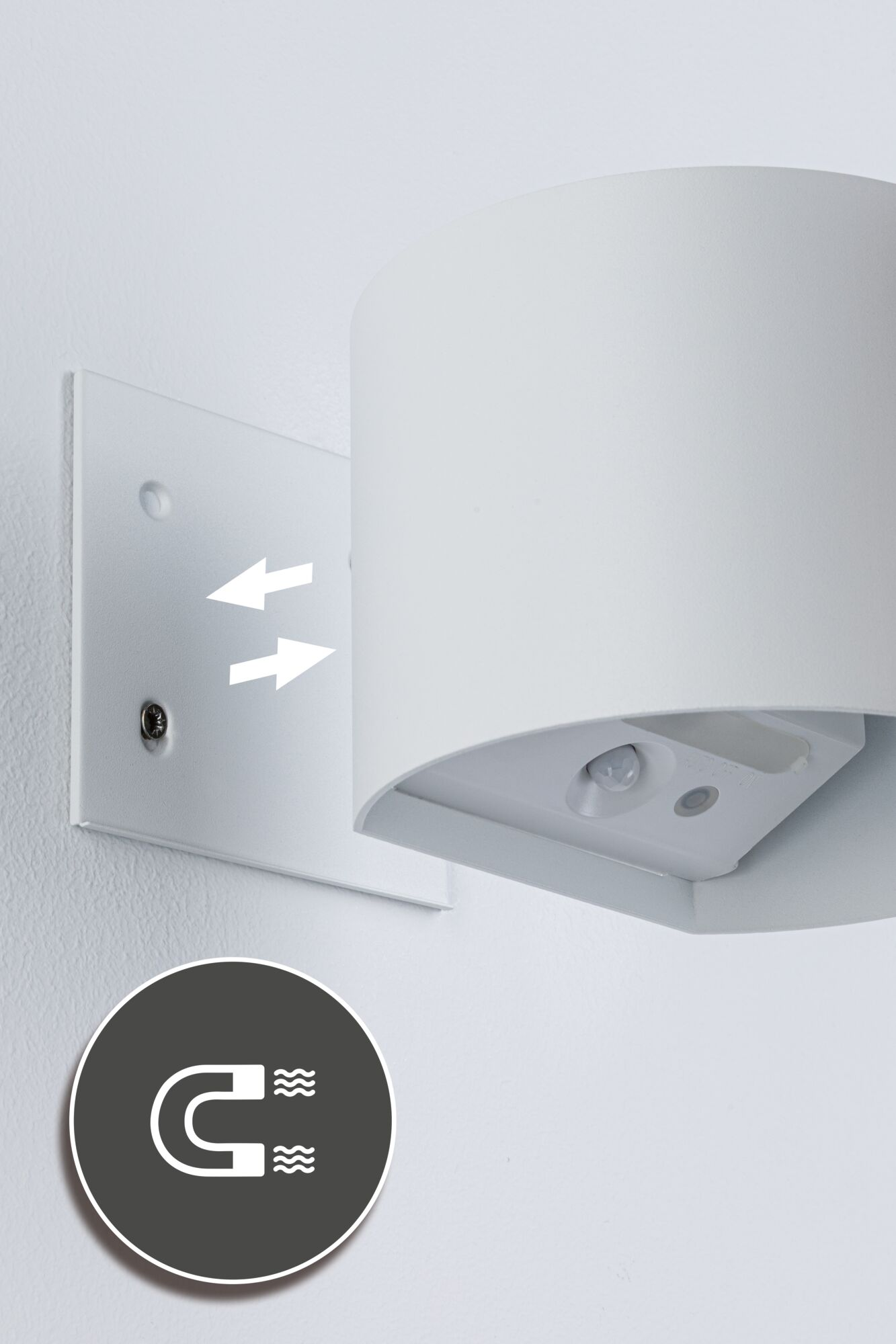 Biały uchwyt ścienny ze strzałkami i ikoną magnesu. Po prawej wisi oprawa oświetleniowa.