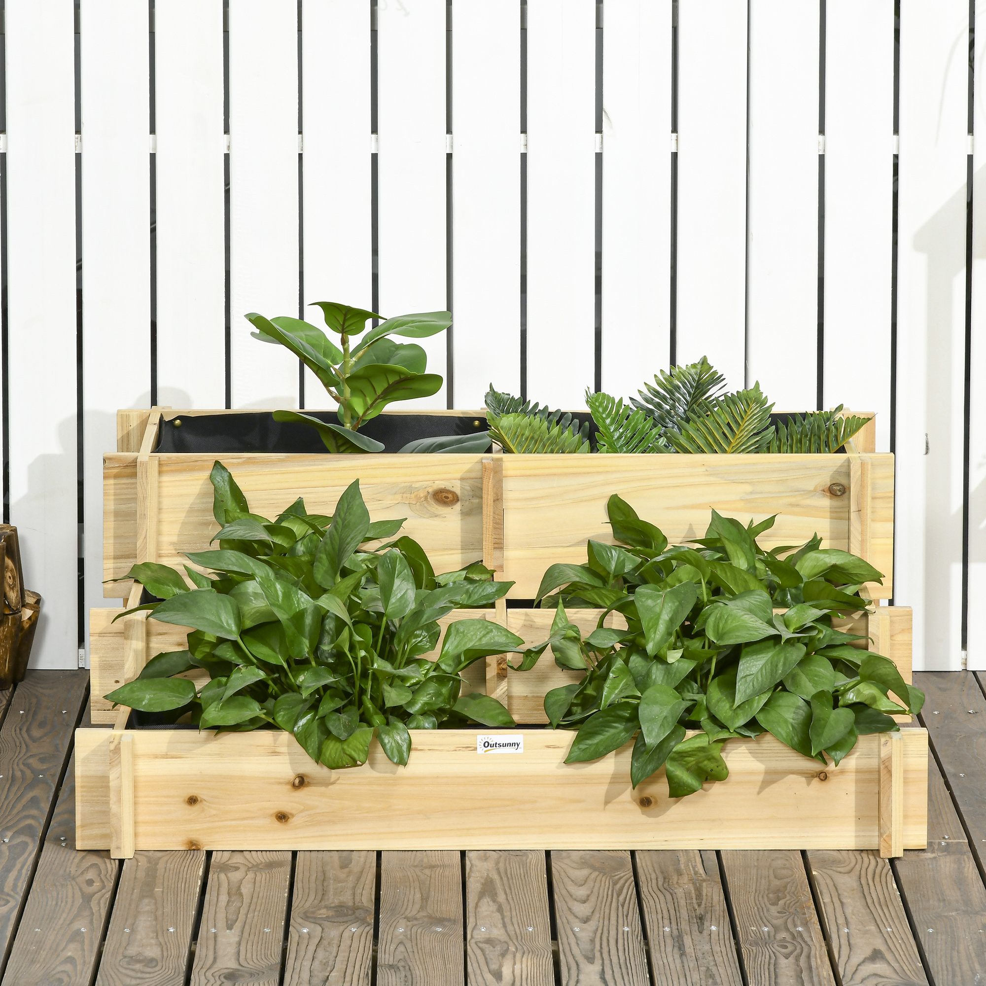 Una jardinera de madera escalonada con exuberantes plantas verdes se asienta sobre una cubierta de madera ante una valla de listones blancos.
