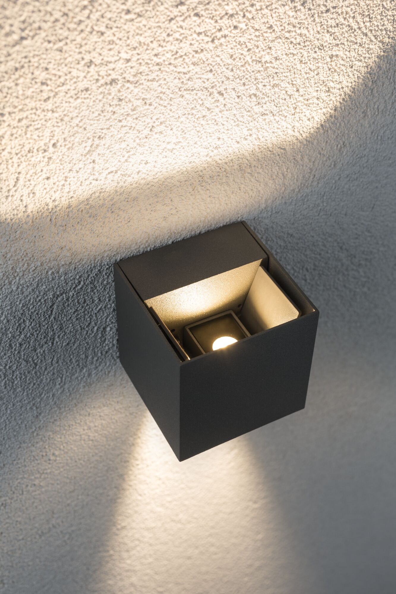 Ciemna, sześcienna lampa zewnętrzna oświetla teksturowaną ścianę ciepłym światłem.