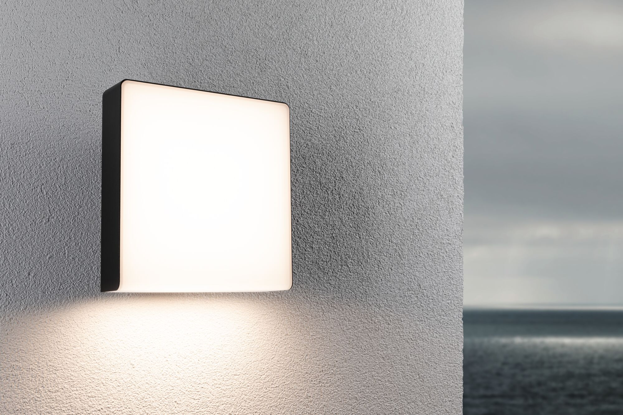 Prostokątna lampa ścienna oświetla szarą ścianę i ocean.