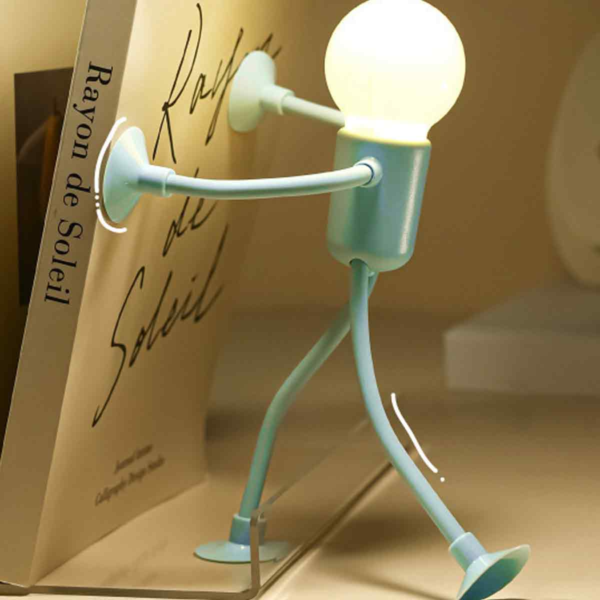 Eine hellblaue Schreibtischlampe in Form einer Figur mit einem Glühbirnenkopf steht neben einem Buch.