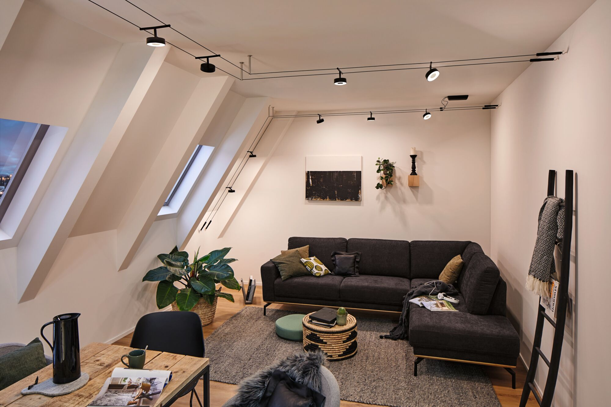 Ein Wohnzimmer mit schrägem Dach, dunklem Sofa, Holztisch und Schienenlichtern.