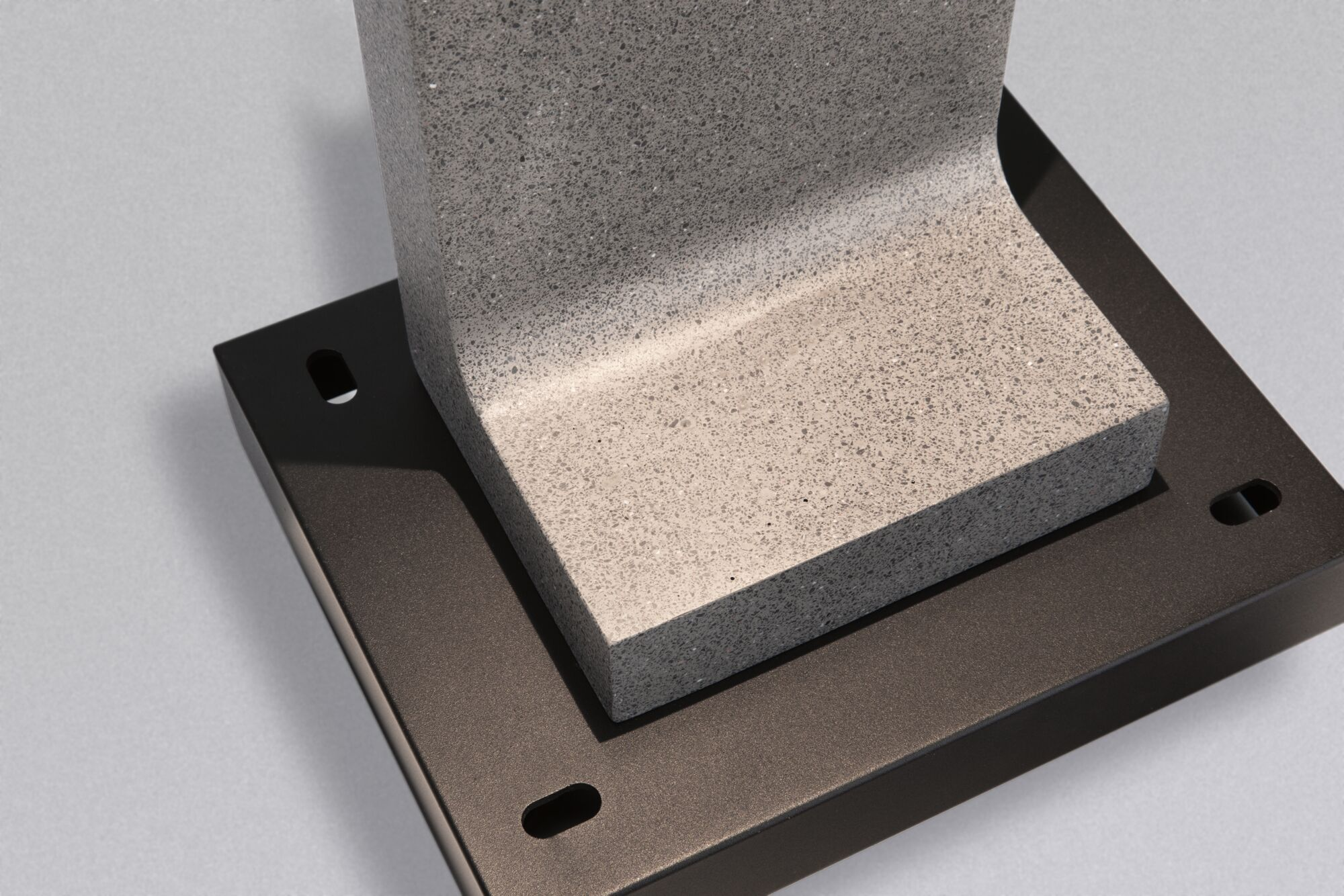 Szary element kamienny na ciemnej metalowej podstawie z widocznymi otworami.