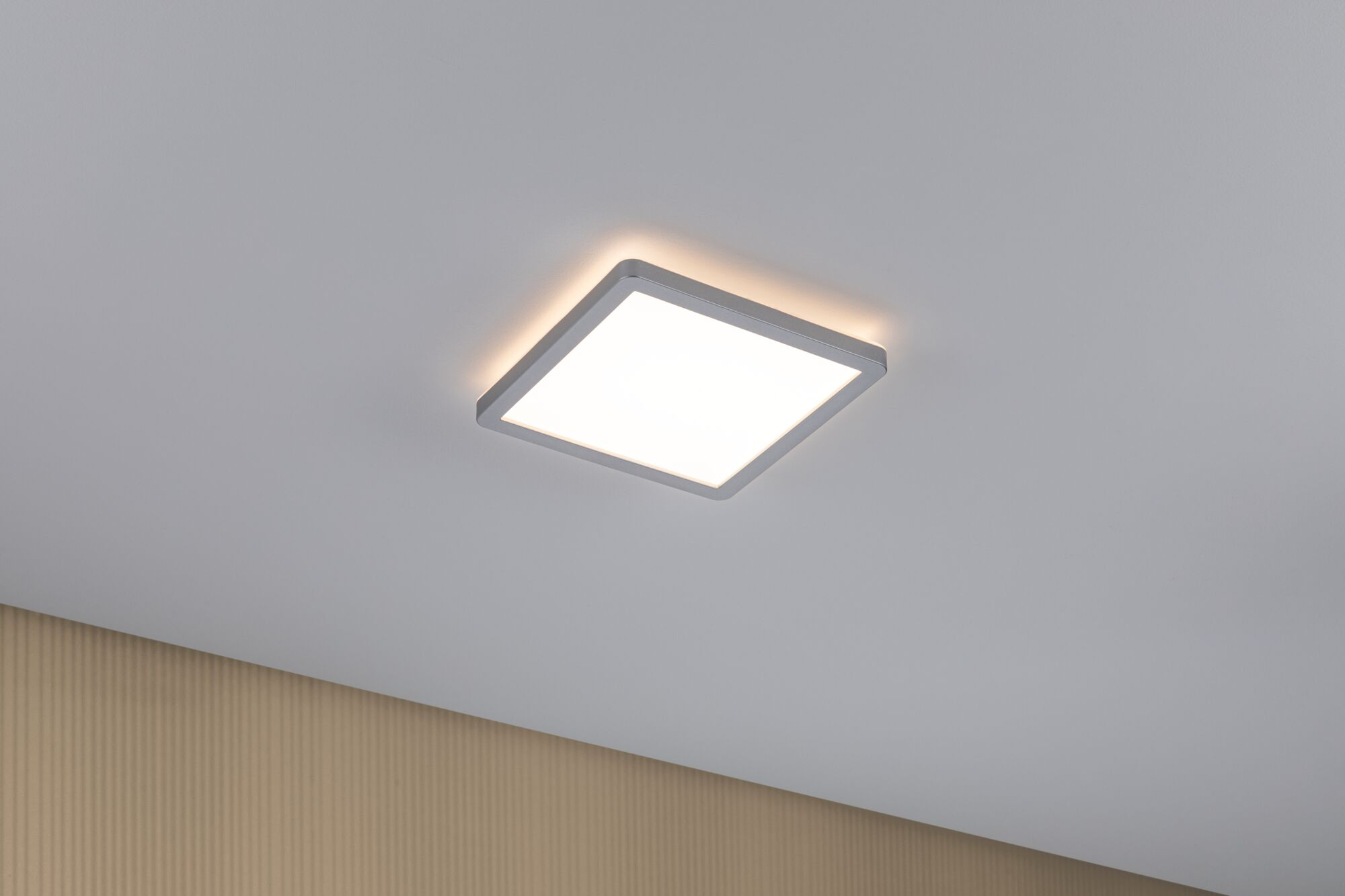 Ein quadratisches Deckenlicht mit silbernem Rahmen leuchtet warmweiß auf einer weißen Decke.