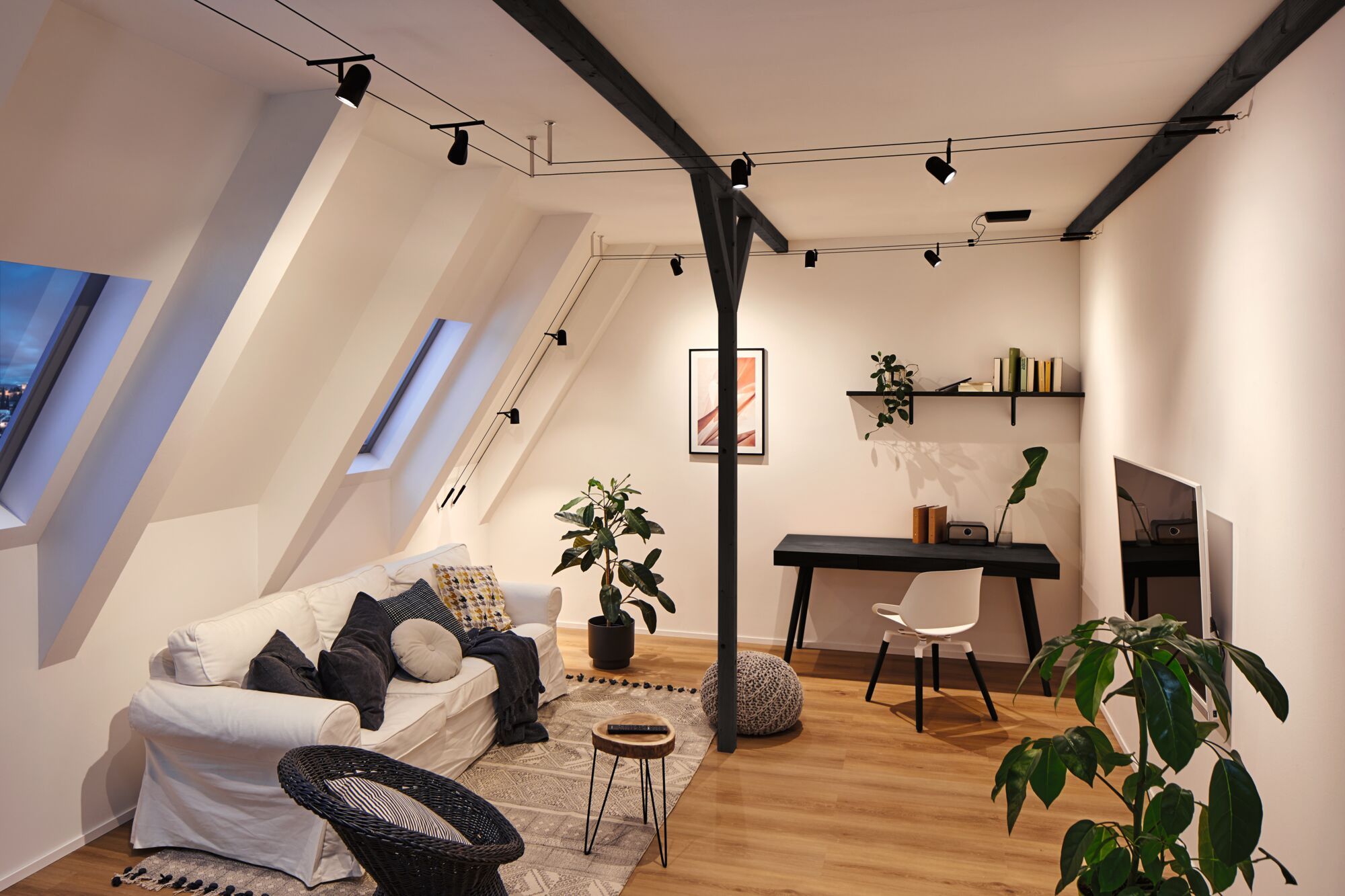 Jasny salon ze skośnymi sufitami, sofą, biurkiem i roślinami.