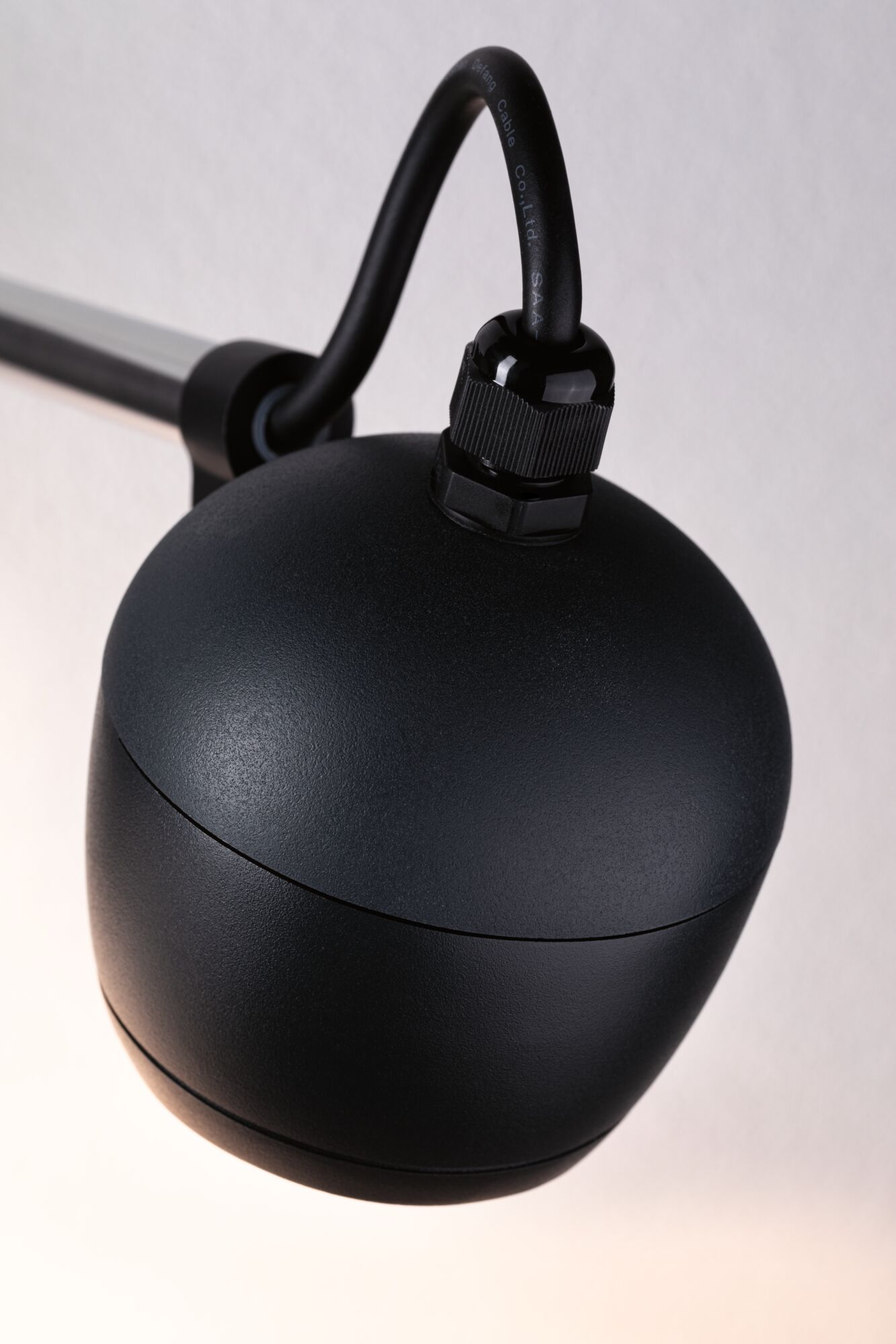 Czarna głowica lampy z przyłączem kablowym i metalowym ramieniem na jasnym tle.