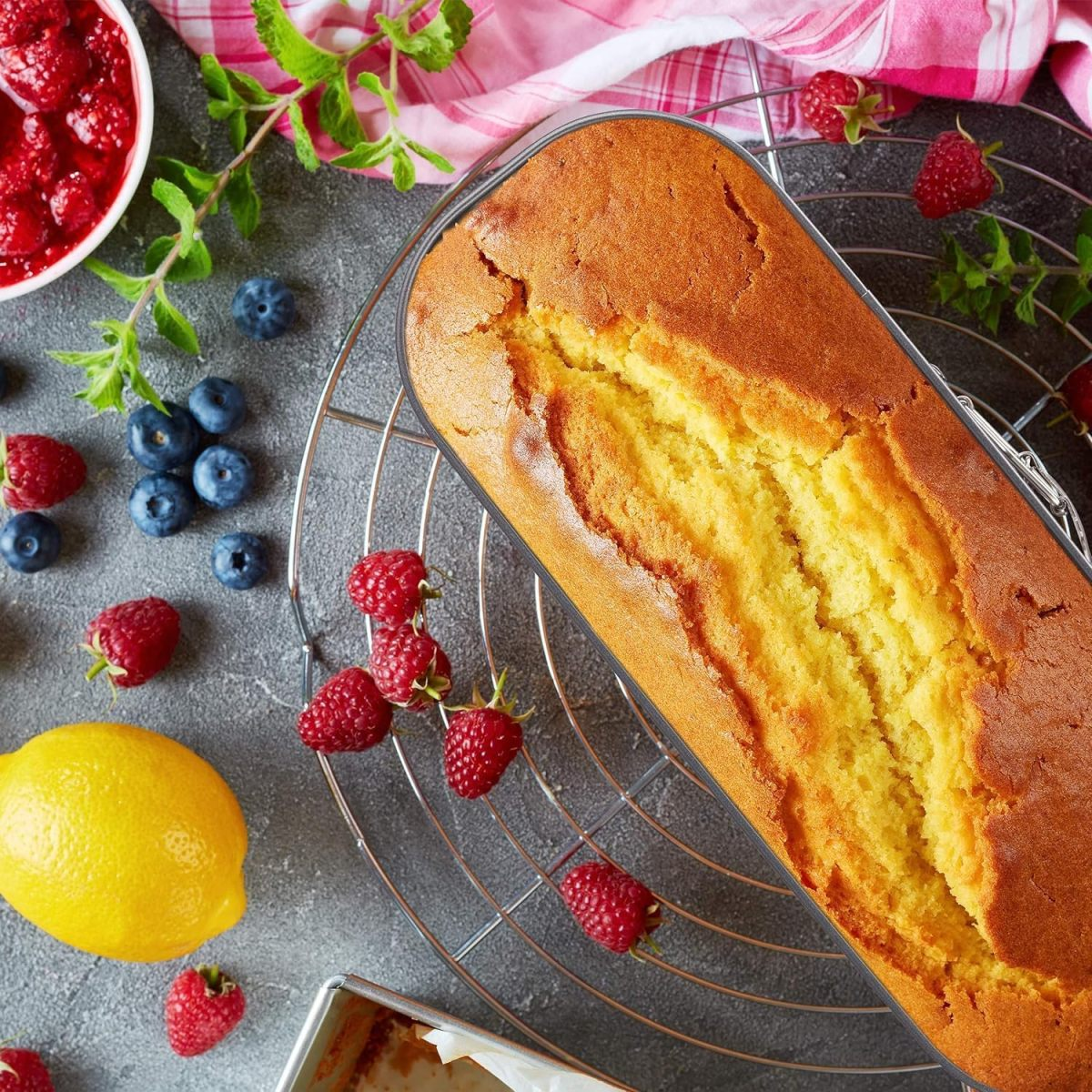 Ein goldbrauner Kuchen in einer Backform, umgeben von Beeren, Zitrone und Minze.