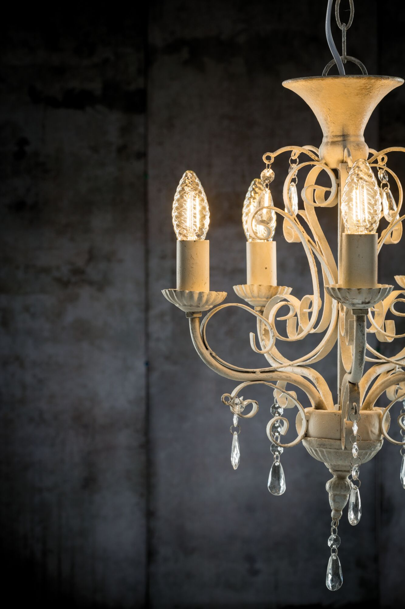 Kremowy żyrandol z kryształowymi wisiorkami wisi na ciemnym tle.