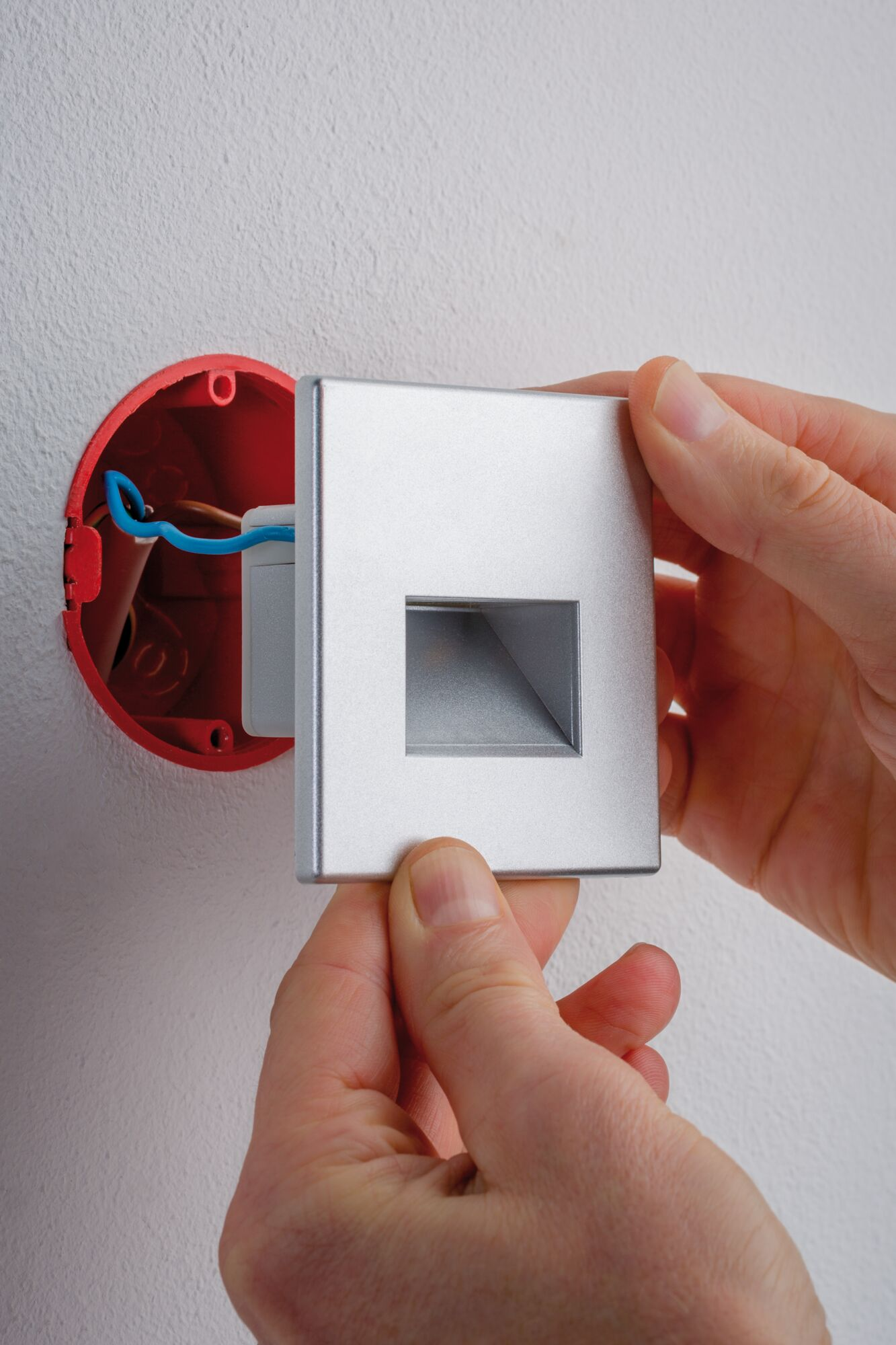 Mains installant un cache-lumière argenté sur une boîte électrique rouge.