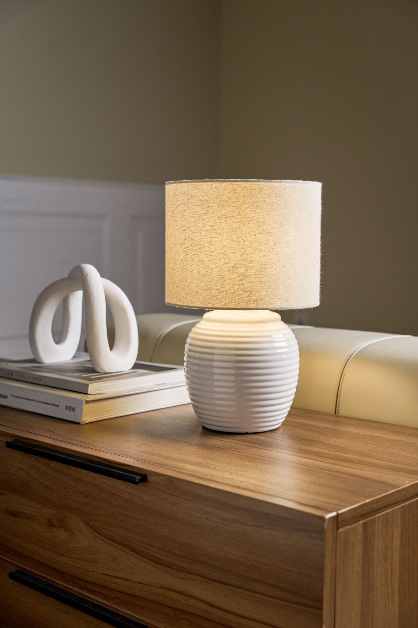 Biała lampa stołowa na drewnianej komodzie obok rzeźby.