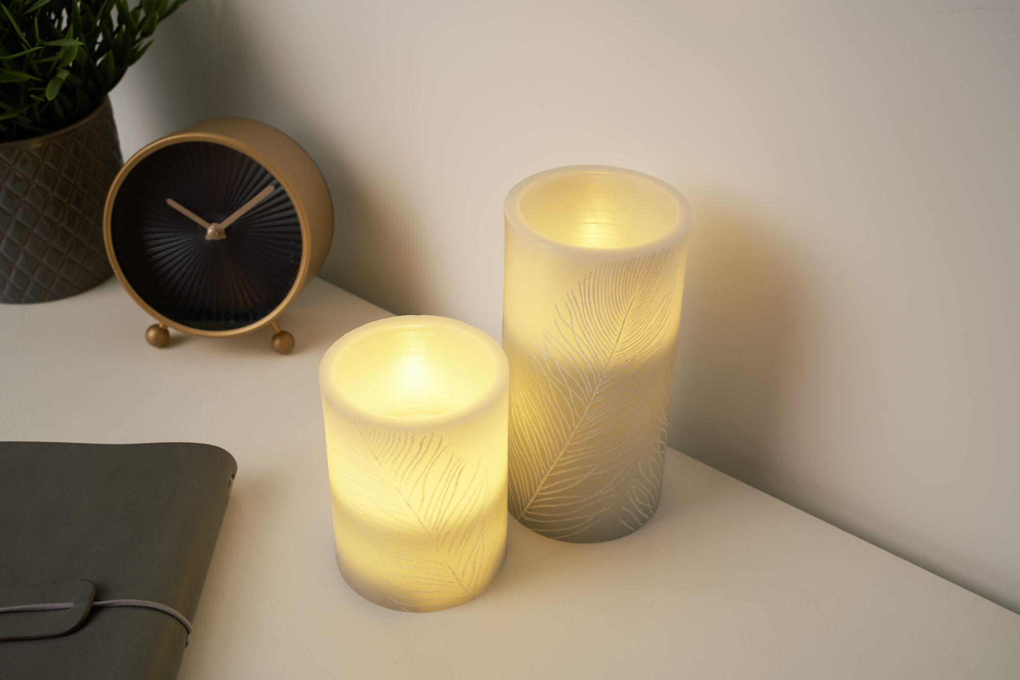 Dwie zapalone świece z motywem piór, zegar i roślina na stole.