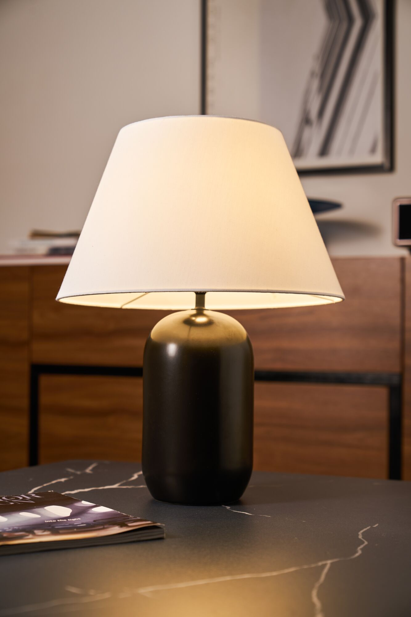 Ciemna lampa stołowa z białym kloszem stoi na marmurowym stole z czasopismem.