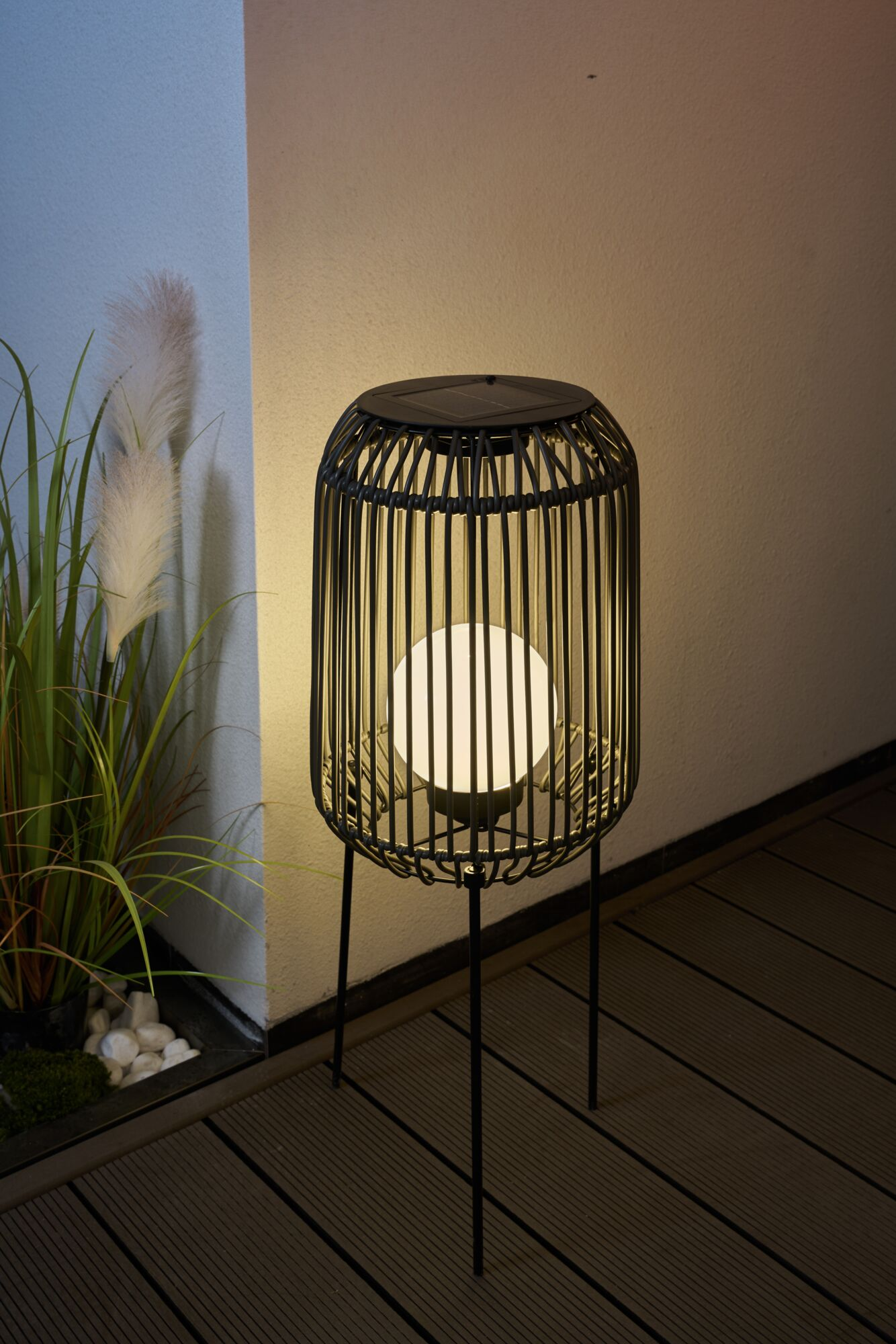 Okrągła lampa z plecionym kloszem stoi na drewnianym tarasie obok roślin.