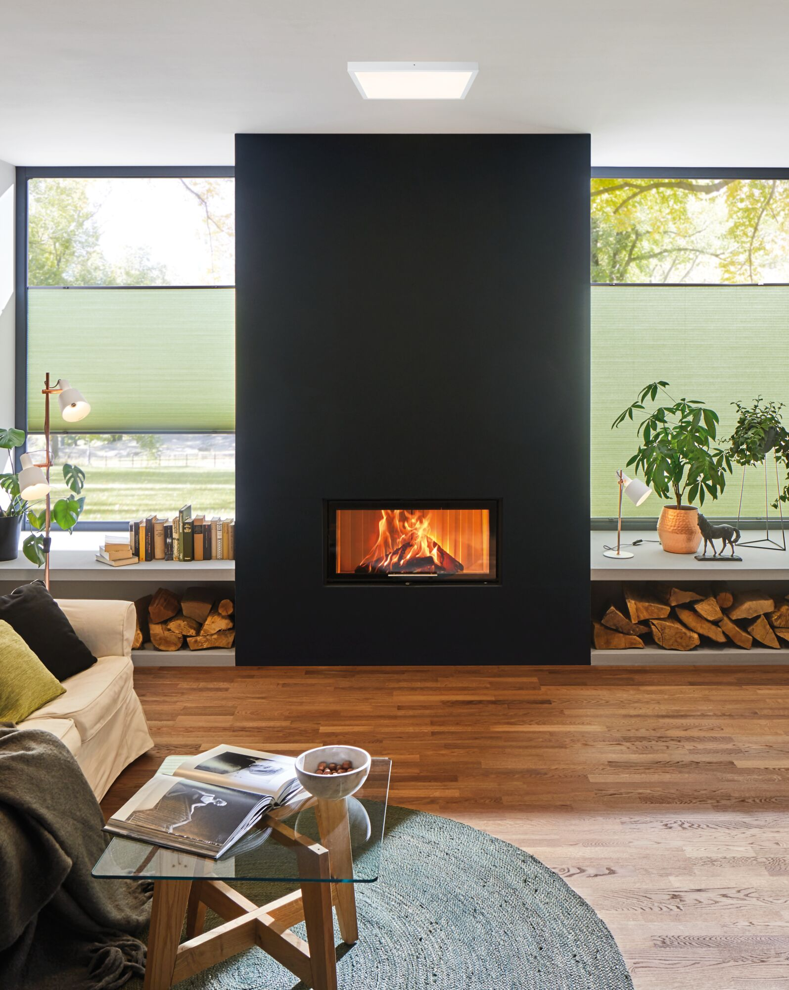 Ein Wohnzimmer mit Kamin, Sofa, Couchtisch und großen Fenstern mit Blick ins Grüne.