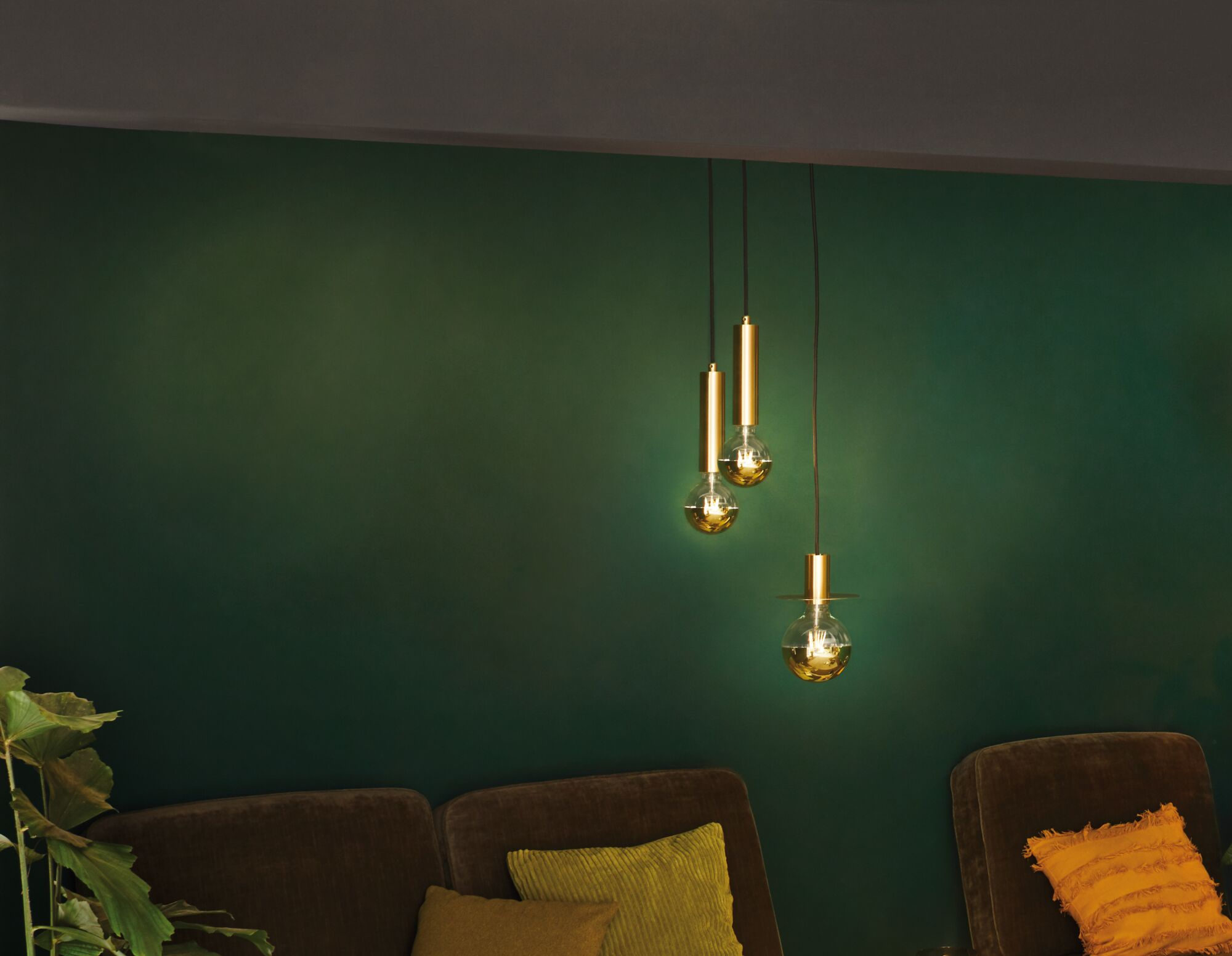 Tre lampade a sospensione dorate con lampadine pendono contro un muro verde scuro.