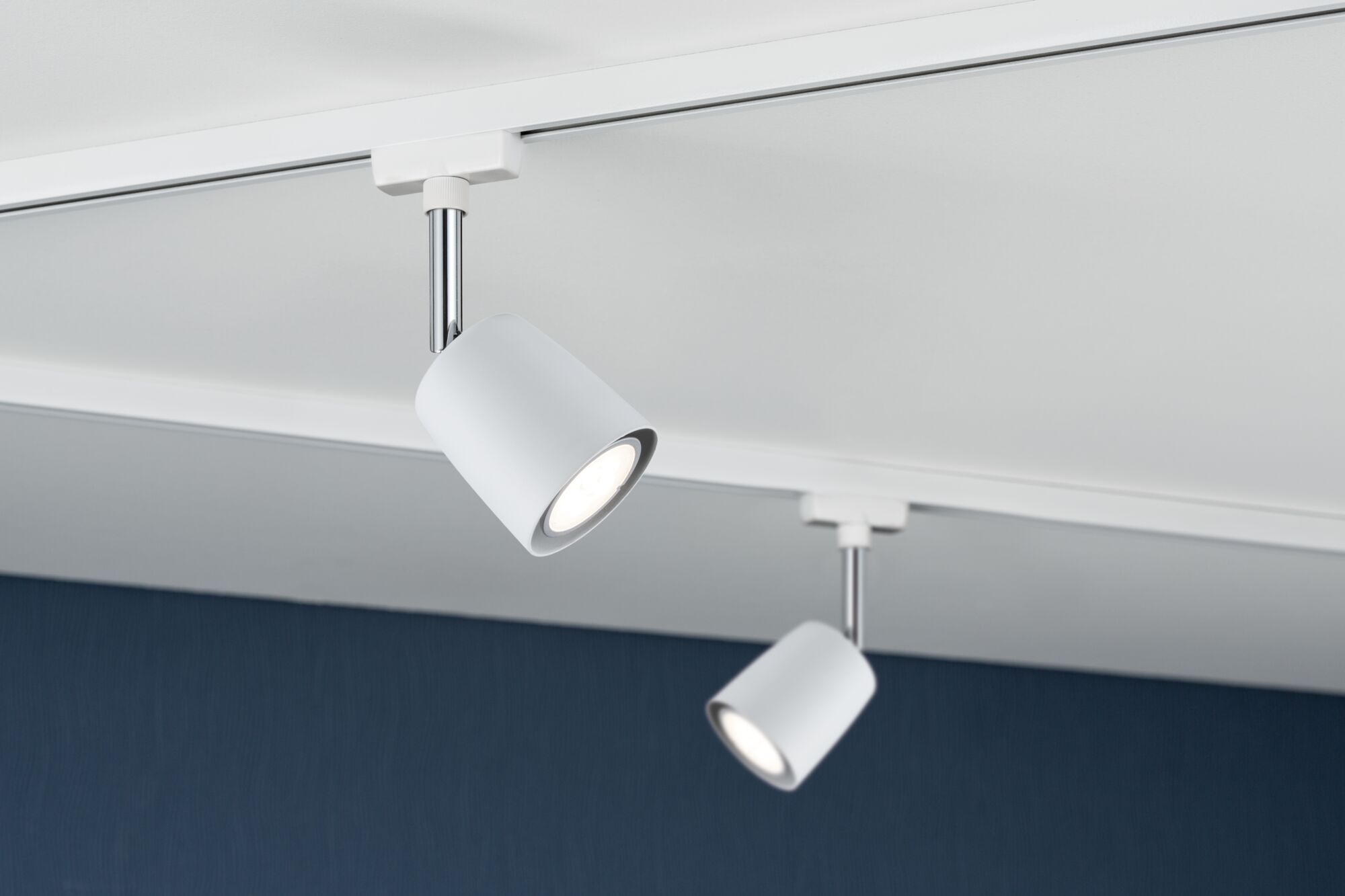Due faretti cilindrici bianchi con accenti cromati su binario bianco contro un muro blu scuro.