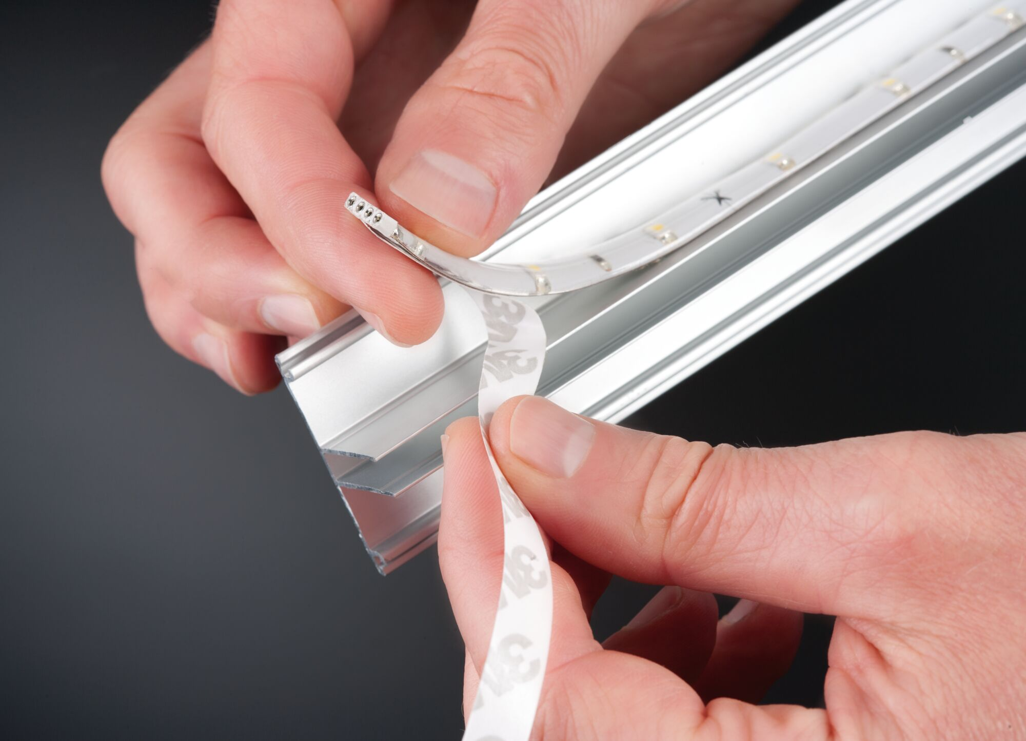 Dłonie zdejmują taśmę z paska LED, by przykleić go do profilu aluminiowego.