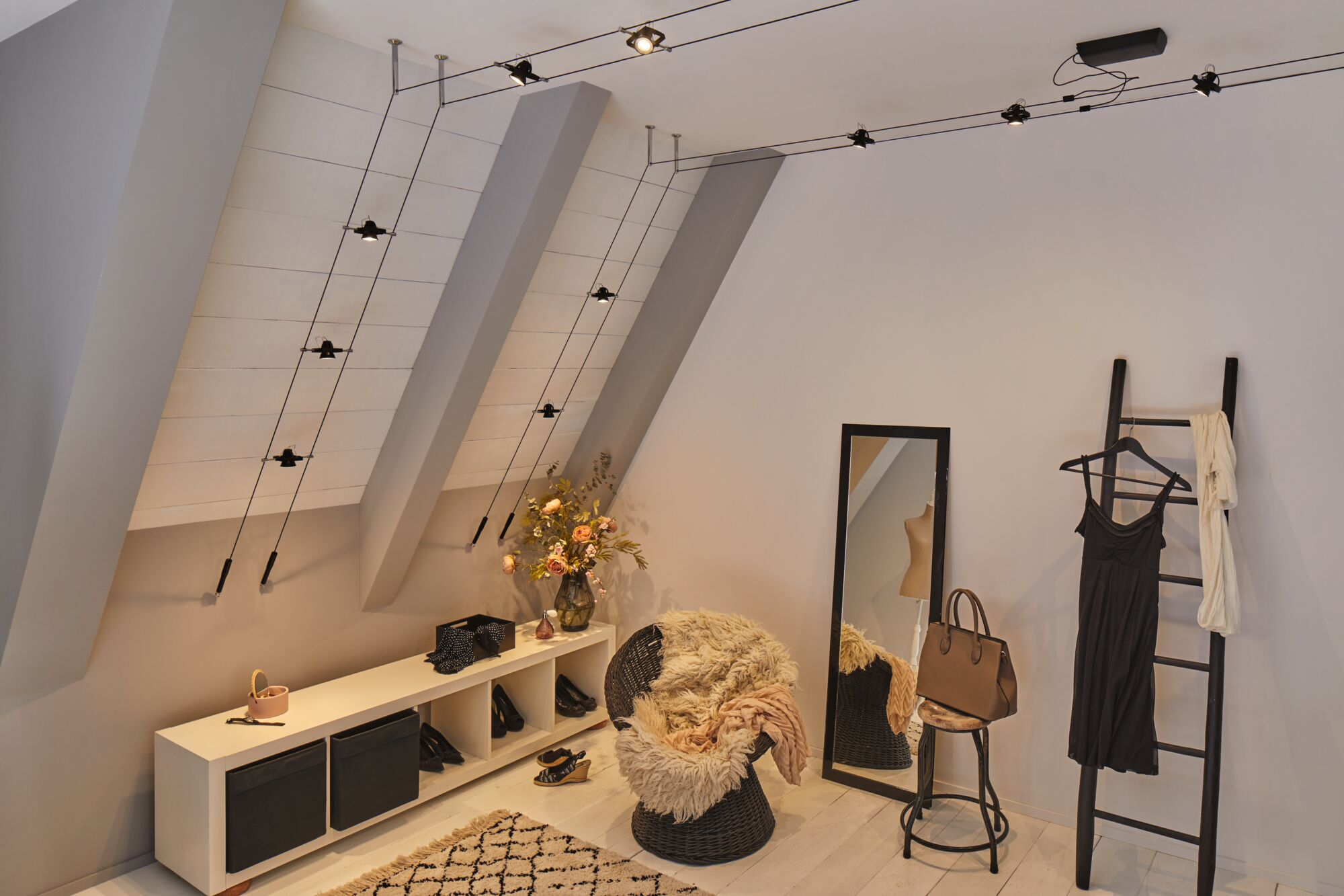 Chambre mansardée avec plafond en pente, éclairage sur rail, étagères, chaise, miroir et échelle avec vêtements.