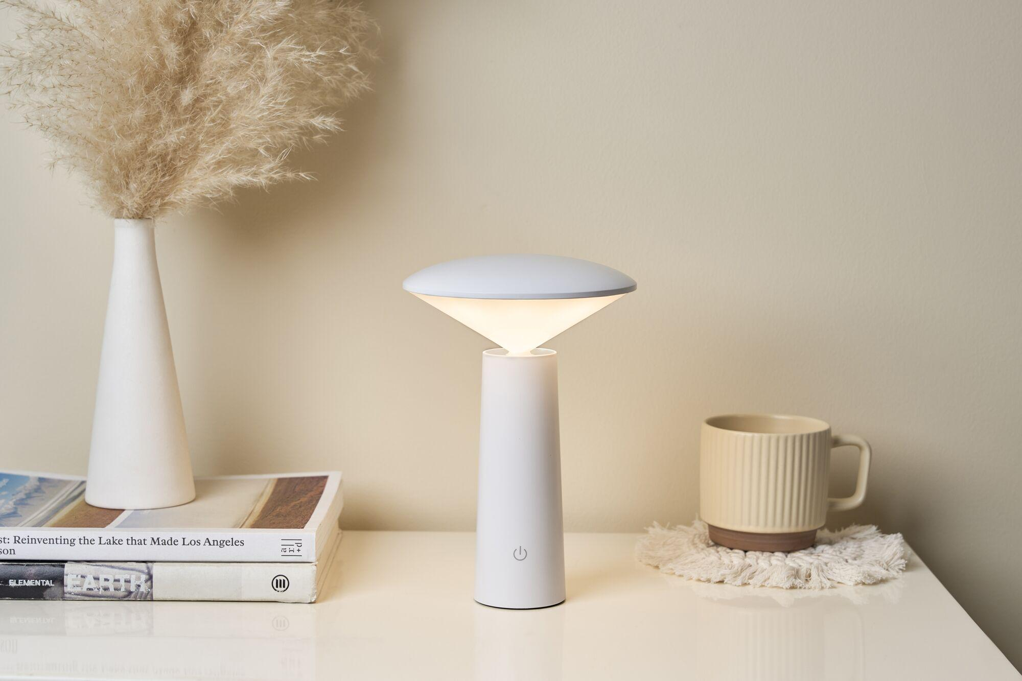 Biały stolik nocny z lampą, książkami, wazonem z trawą pampasową i kubkiem.