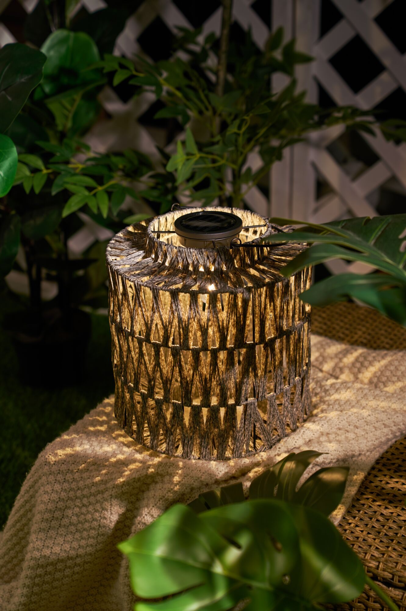 Pleciona latarnia z panelem słonecznym stoi na kocu, otoczona roślinami.