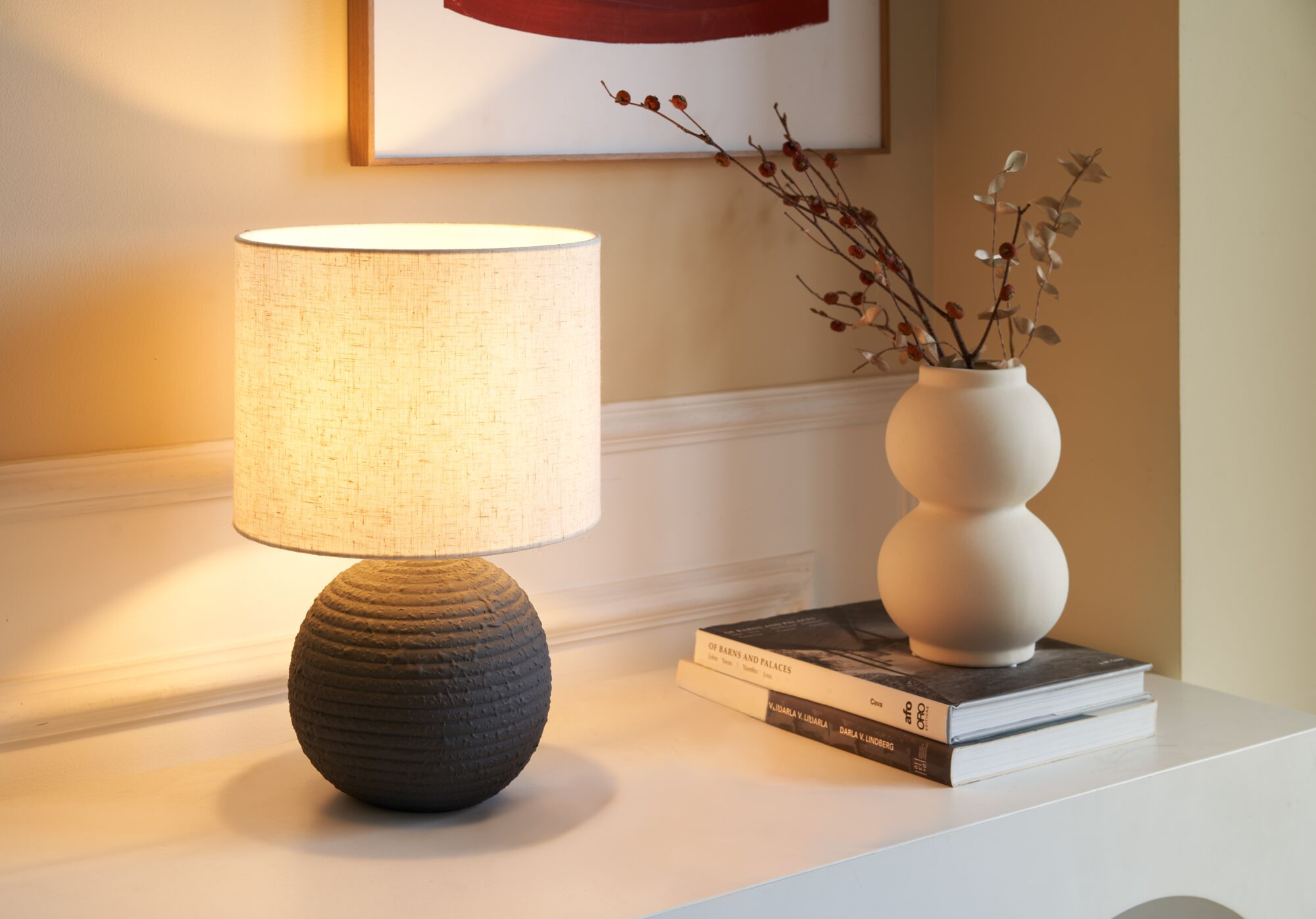 Ciemna, teksturowana lampa stoi na białej powierzchni obok wazonu z suszonymi kwiatami i książek.