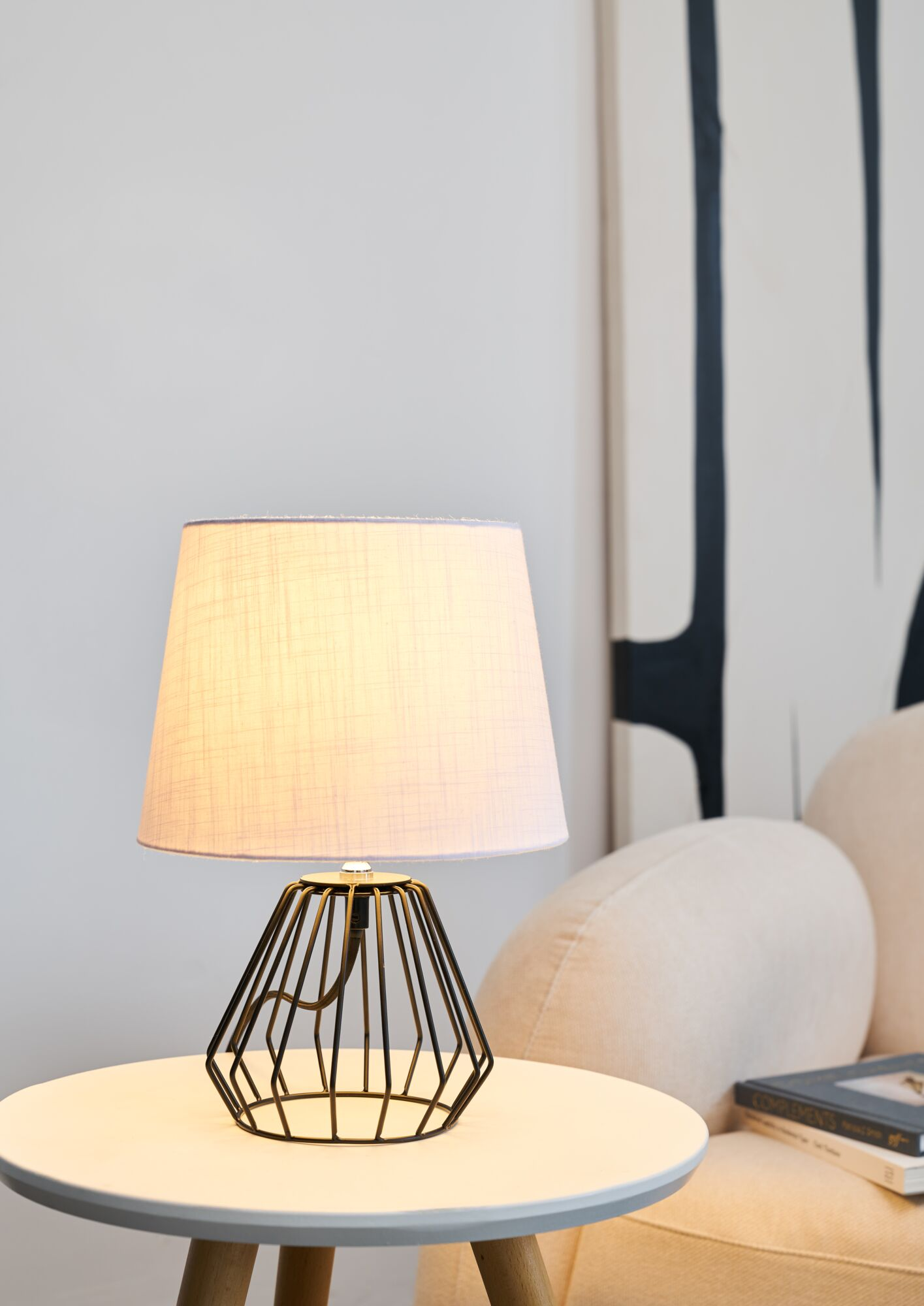Lampka nocna z czarną drucianą podstawą i jasnym kloszem stoi na okrągłym stole.