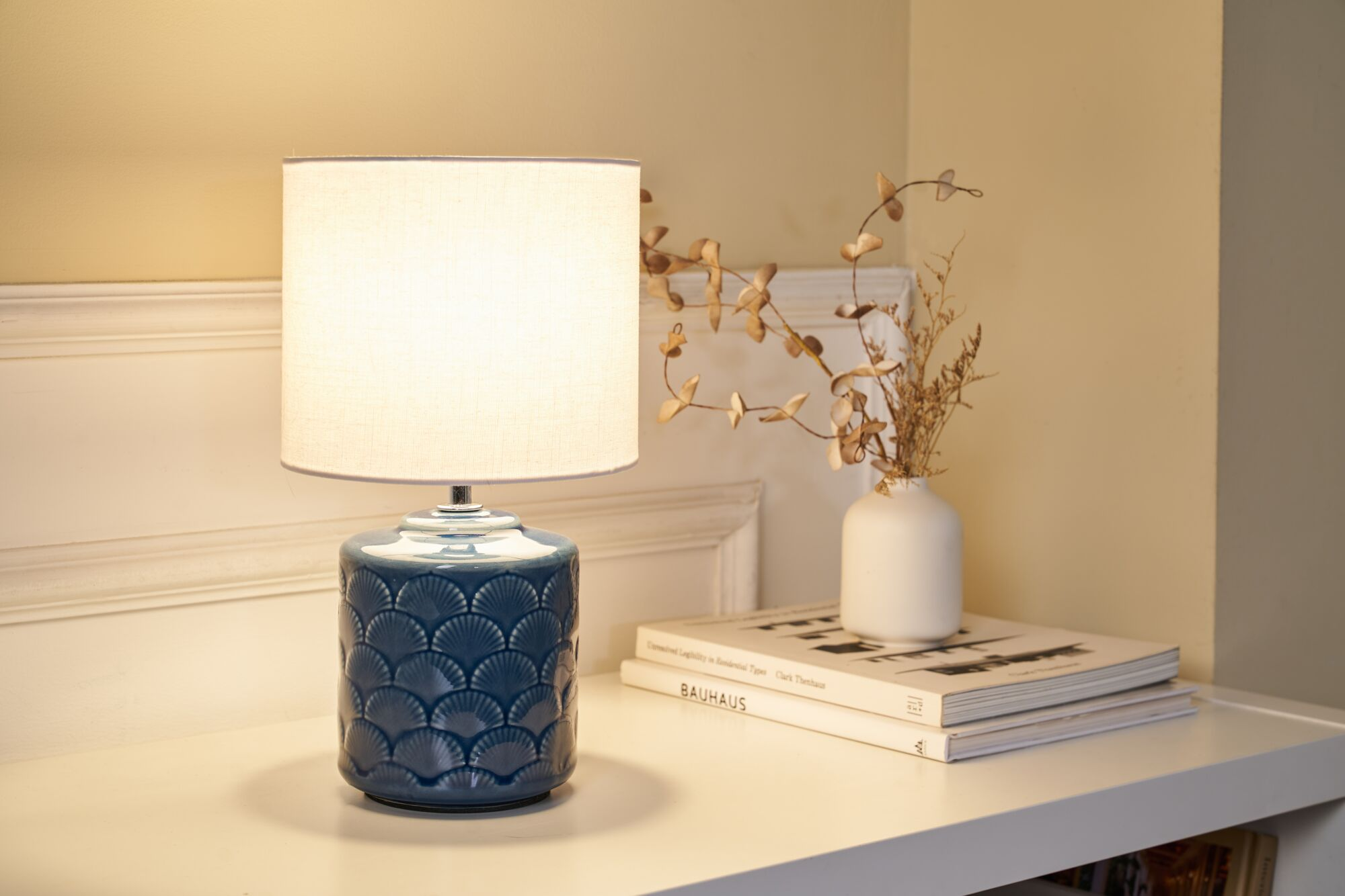 Niebieska ceramiczna lampa z teksturowaną podstawą stoi na półce obok książek i wazonu z suszonymi kwiatami.