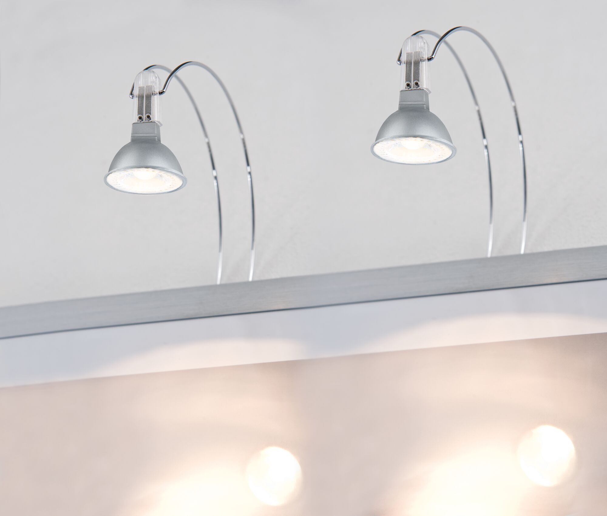 Dwa srebrne reflektory szynowe z zakrzywionymi ramionami oświetlają w dół.