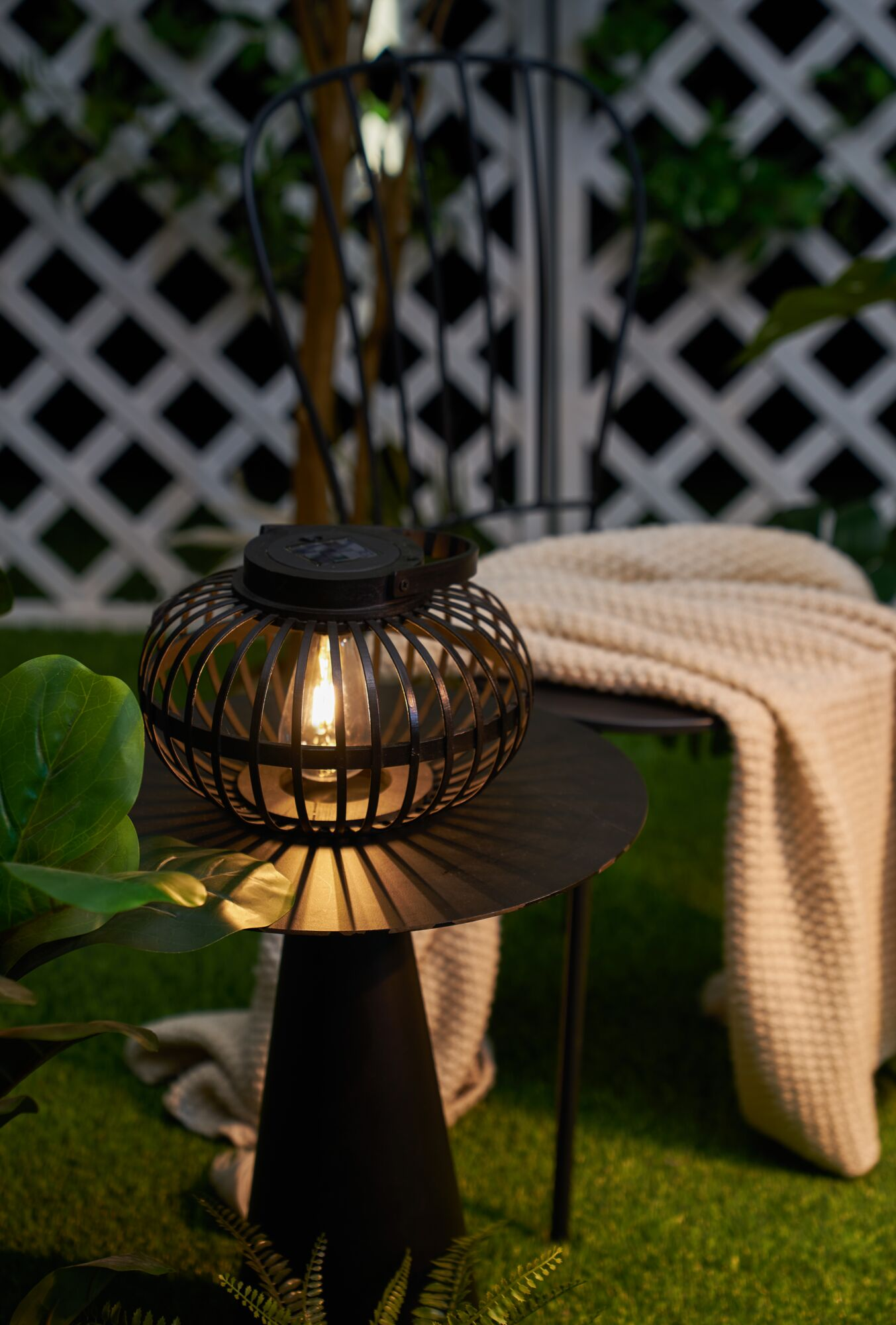 Oświetlony stolik z latarnią na sztucznej trawie z kocem i krzesłem.