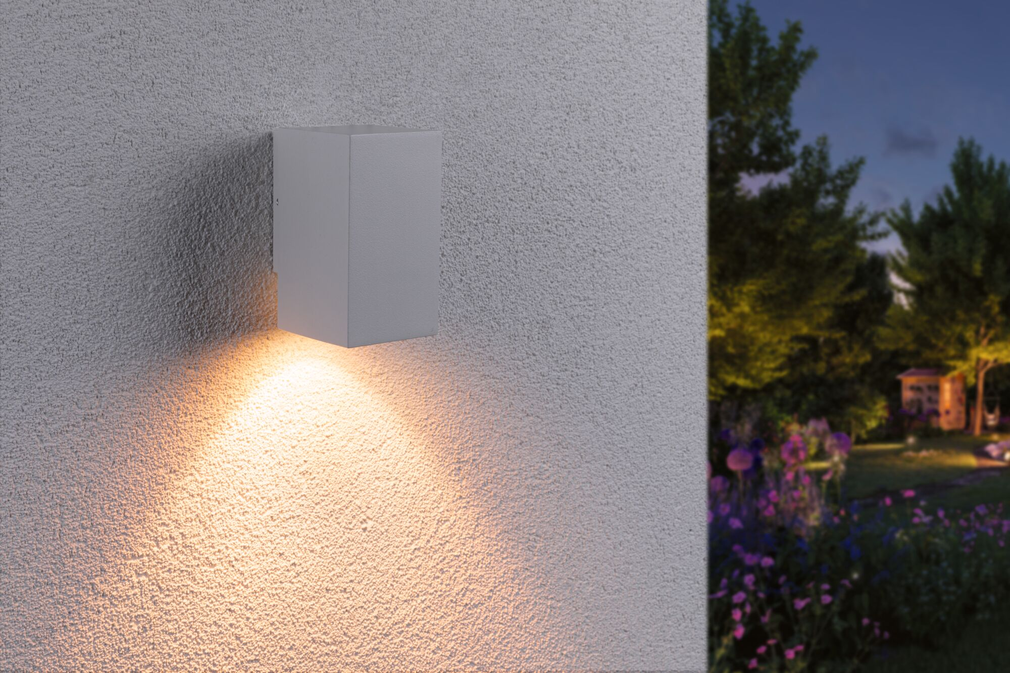 Eine weiße Wandleuchte beleuchtet eine strukturierte Wand. Im Hintergrund ein Garten mit Blumen.