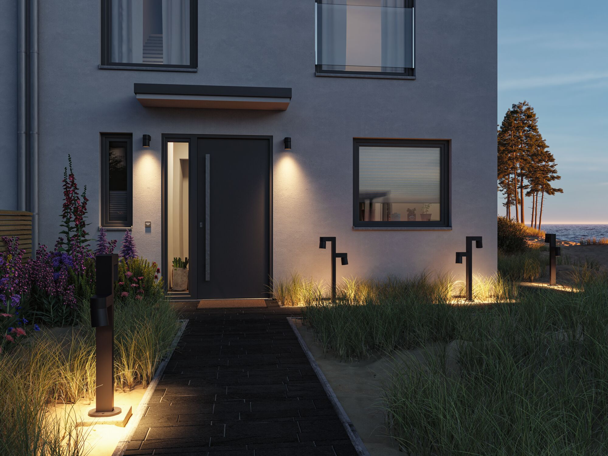 Nowoczesny dom o zmierzchu z oświetloną ścieżką, kwiatami i drzewami nad morzem.